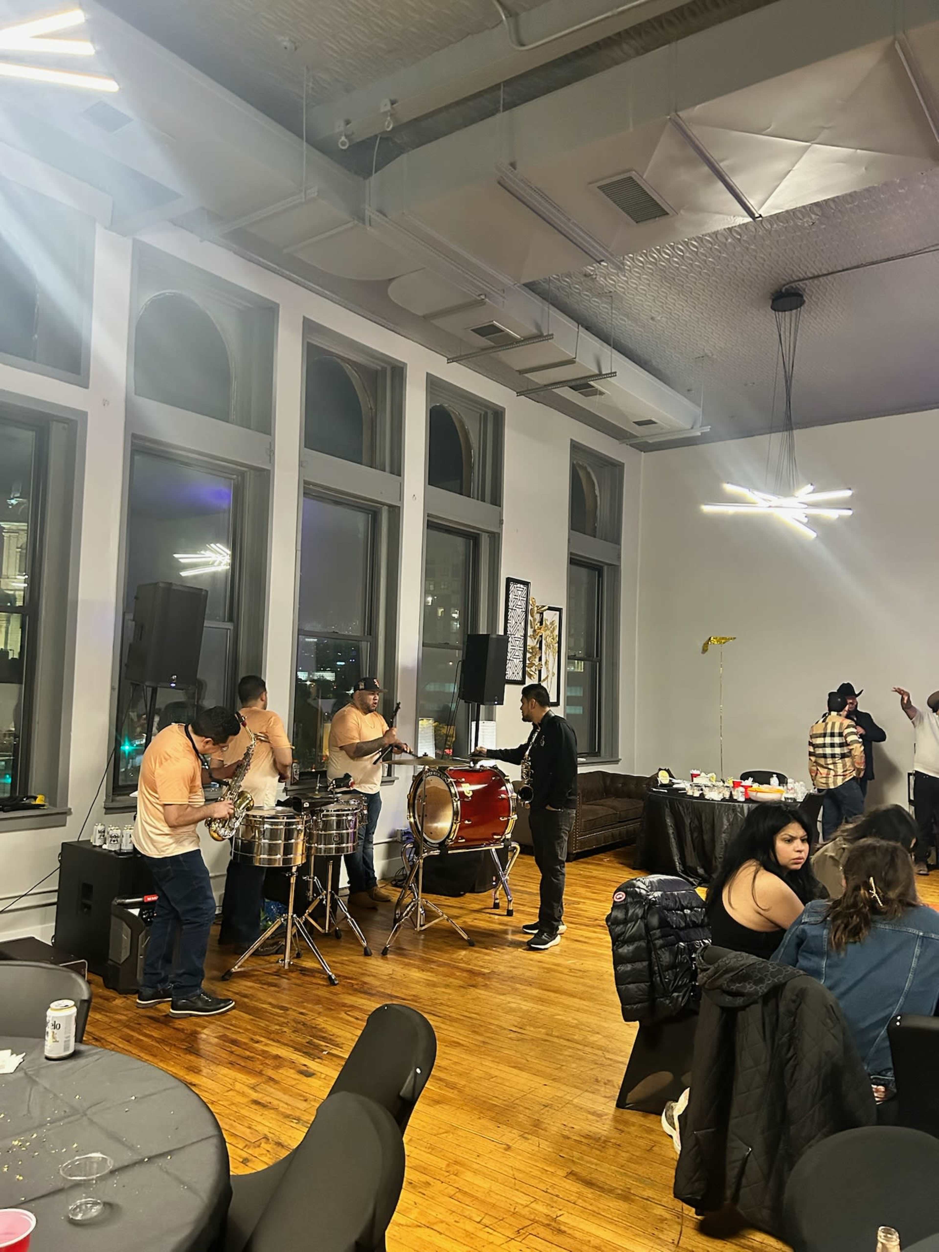 A group of musicians plays drums in a well-lit event space with large windows, while attendees socialize at tables.