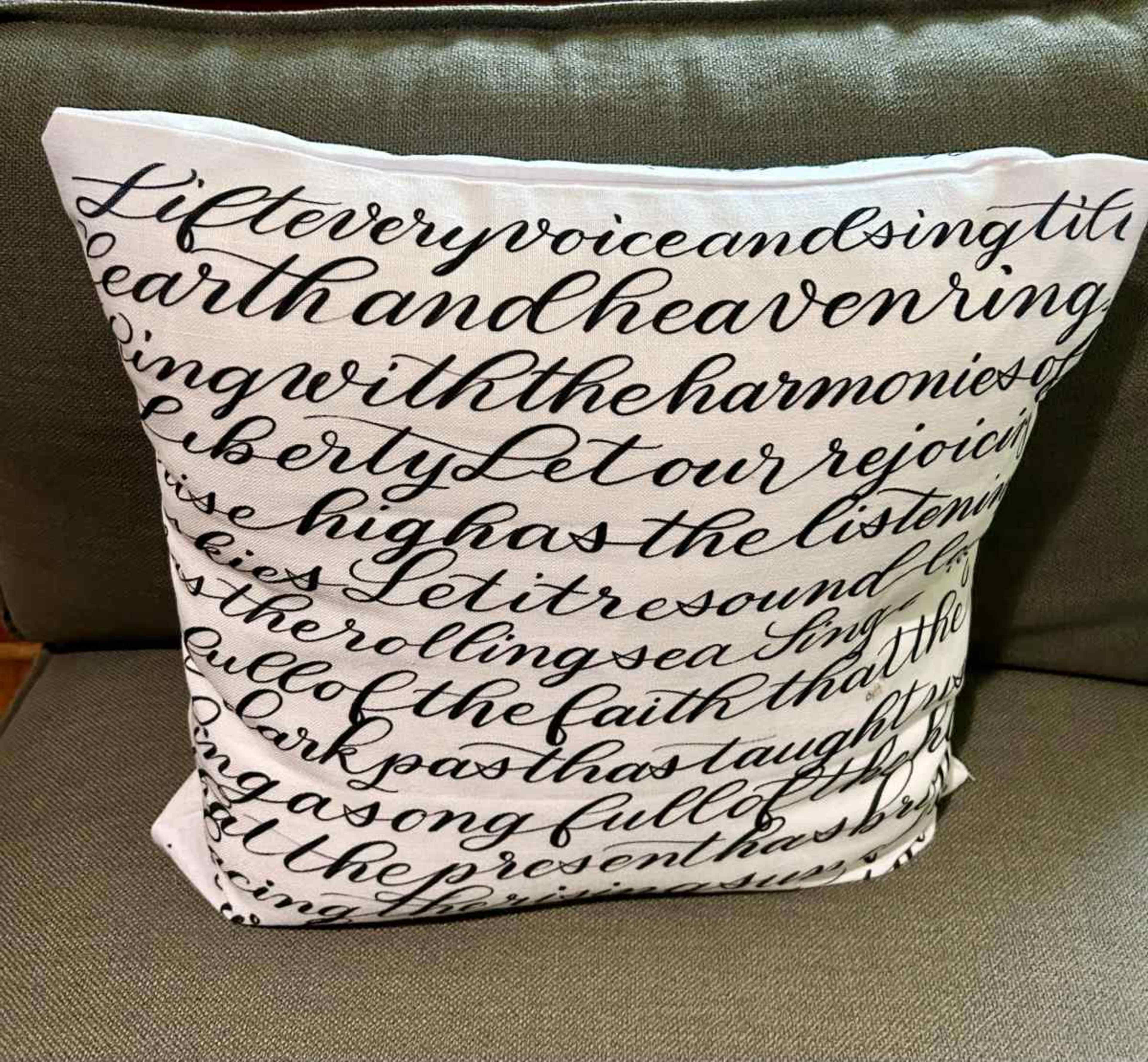The image shows a decorative throw pillow with text written in a cursive font on a light background, resting on a couch.