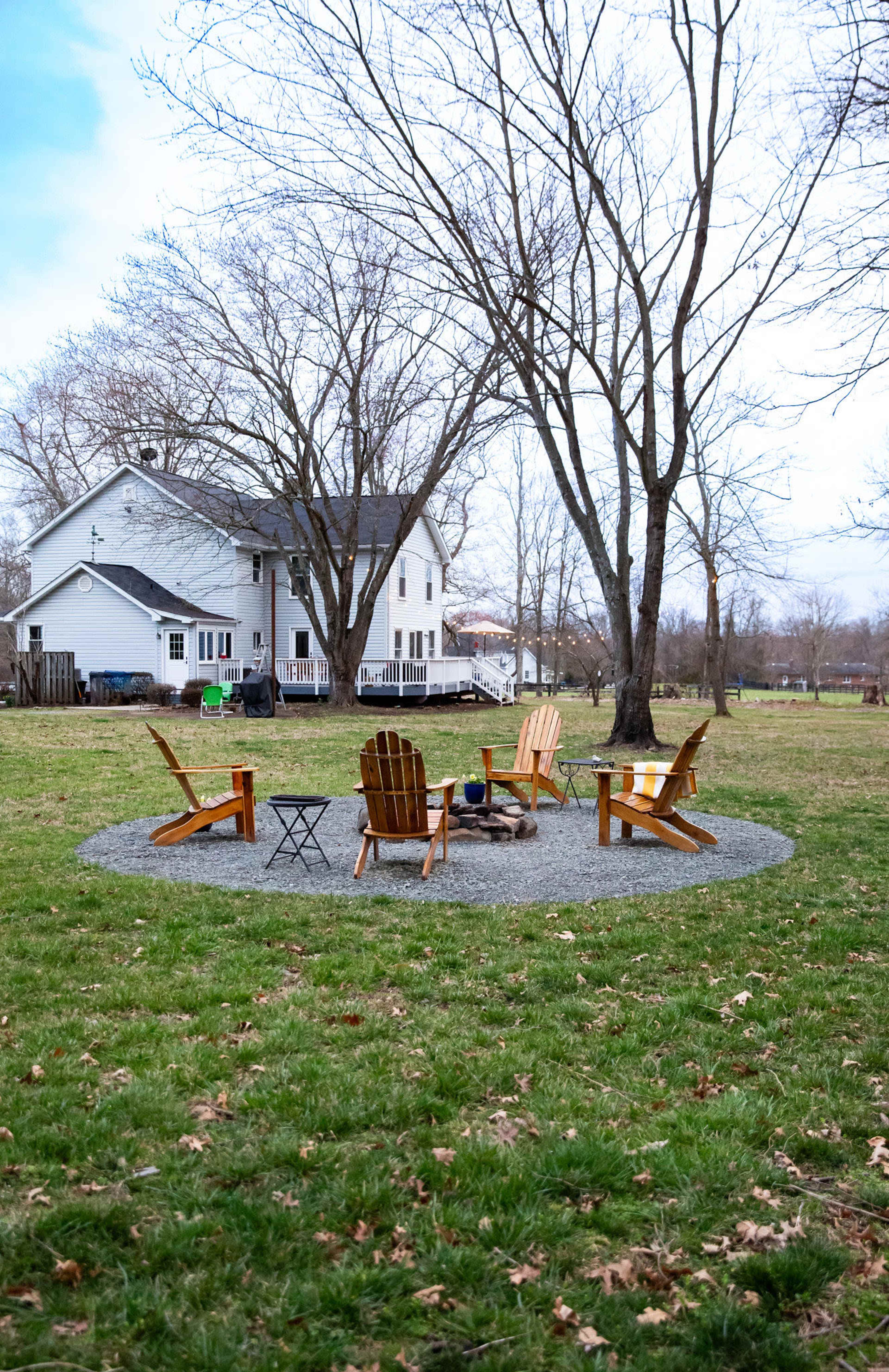 A circular gravel area with four wooden Adirondack chairs surrounding a fire pit is situated in a grassy yard, with a white house and bare trees in the background.