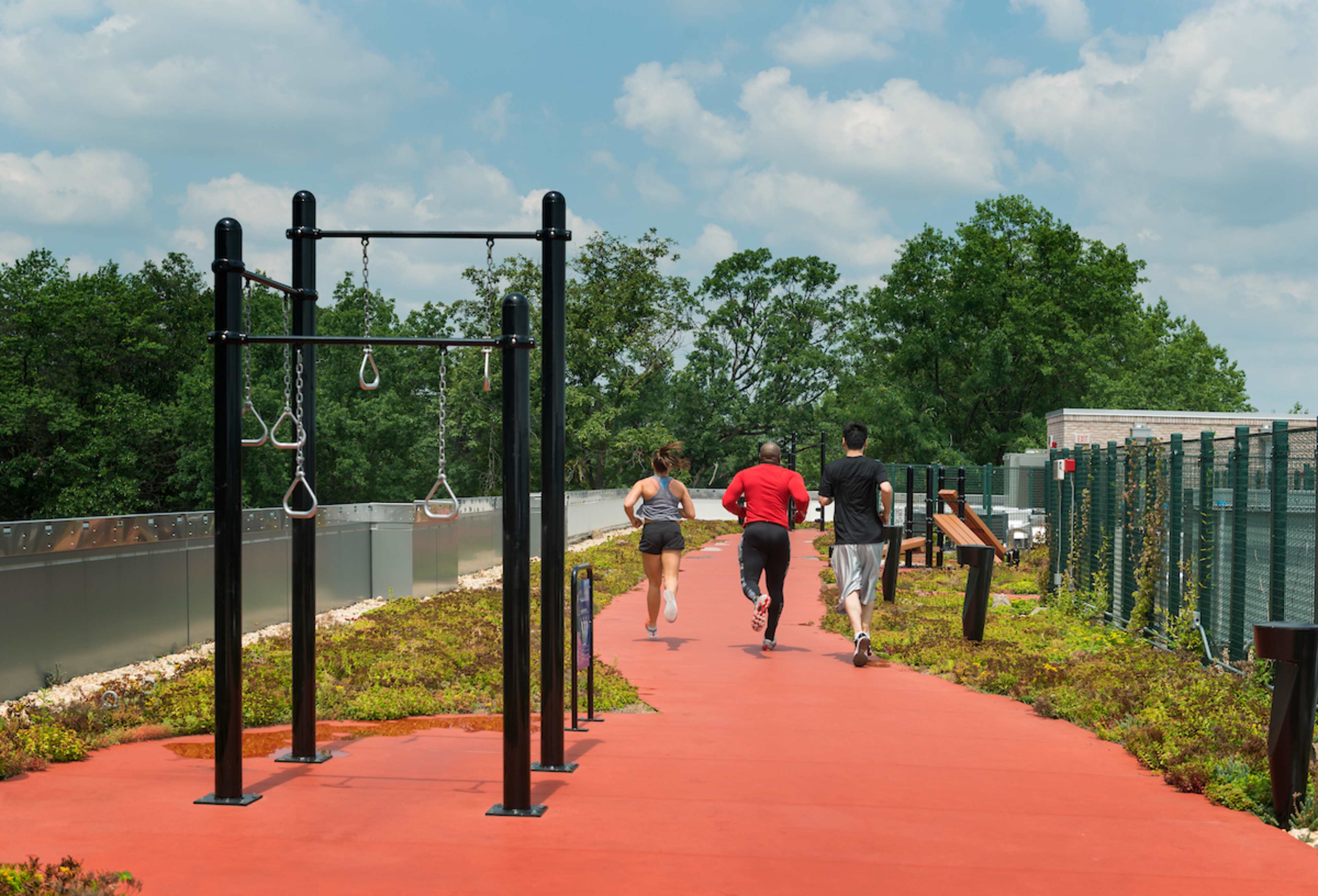 Three people jog down a rubberized path beside fitness equipment in a green outdoor setting.
