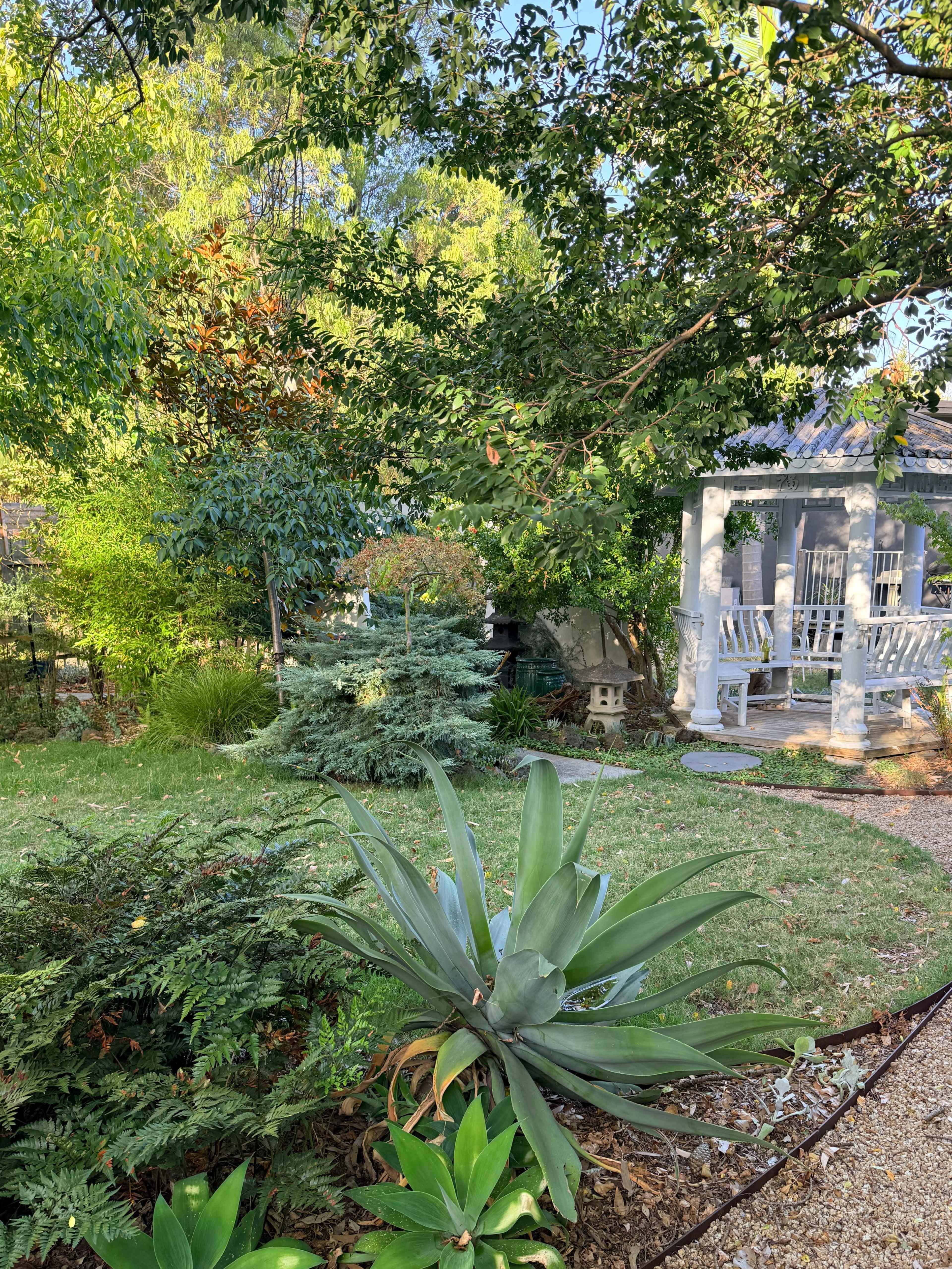 The image shows a landscaped garden featuring various plants, a small gazebo, and a pathway made of gravel.