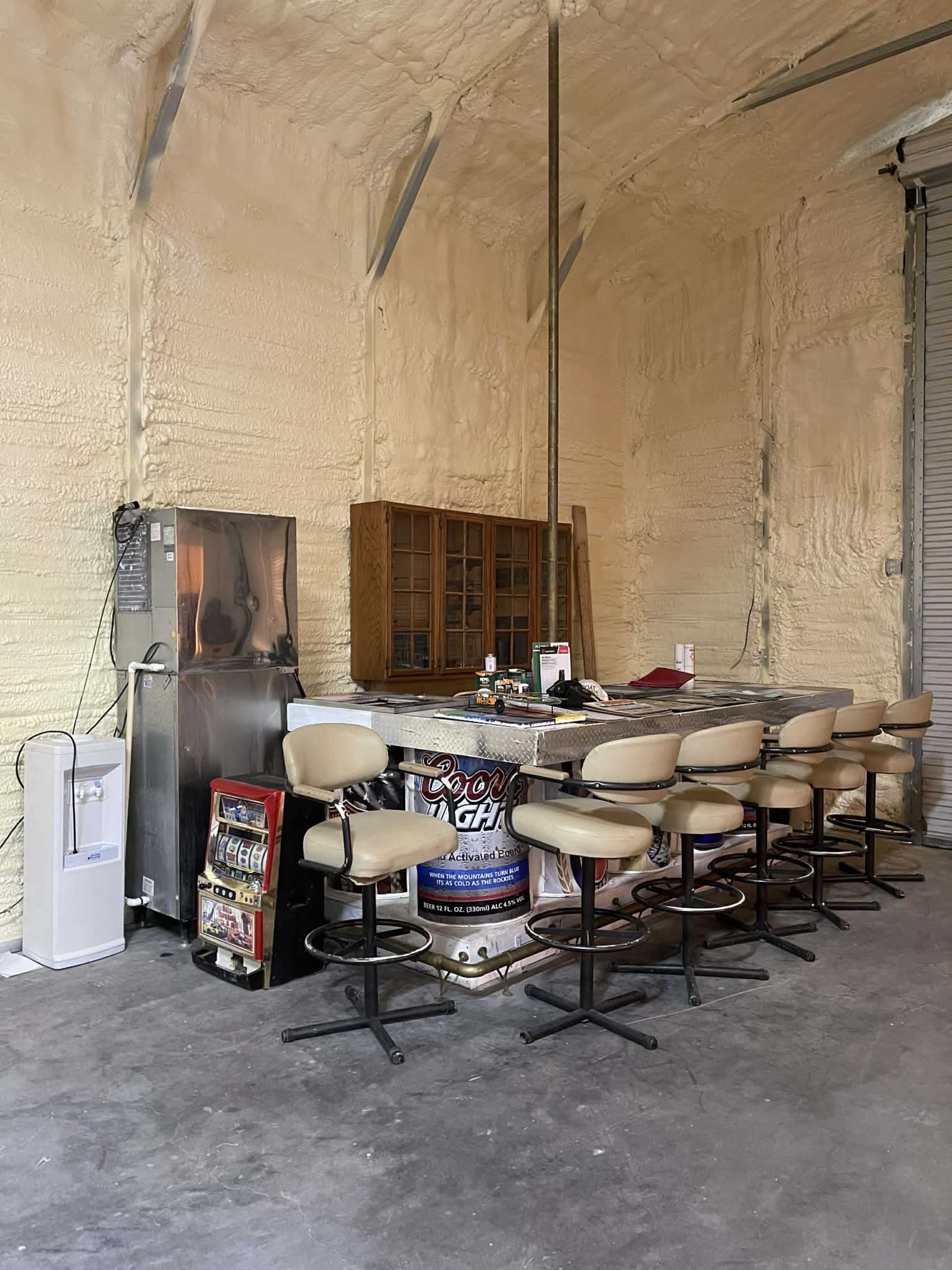 The image shows a bar area with high stools and a counter, surrounded by walls that have a rough, textured finish, and includes a refrigerator and a small vending machine.