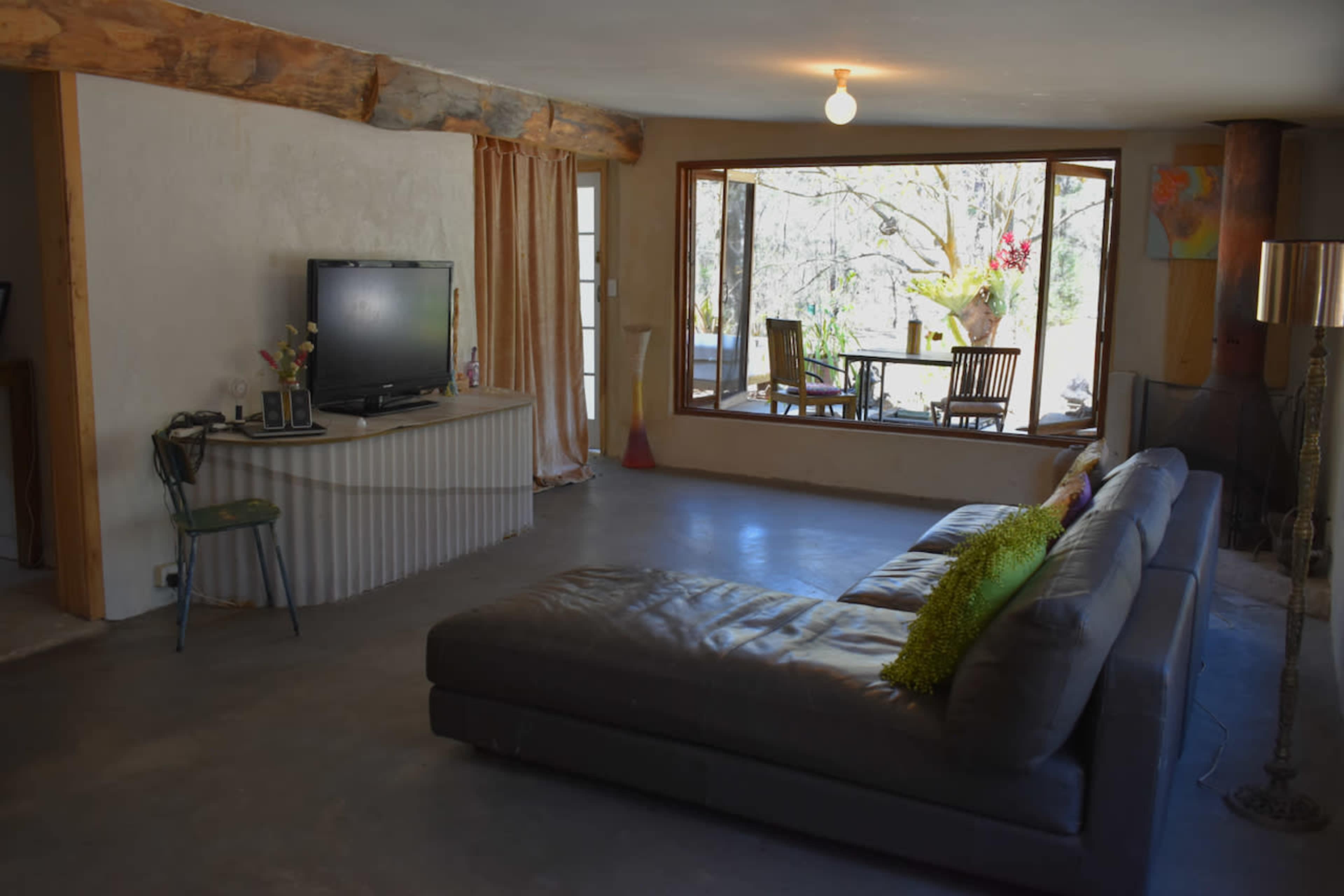 The living room features a large sectional sofa, a TV on a low cabinet, and a large window overlooking an outdoor seating area.