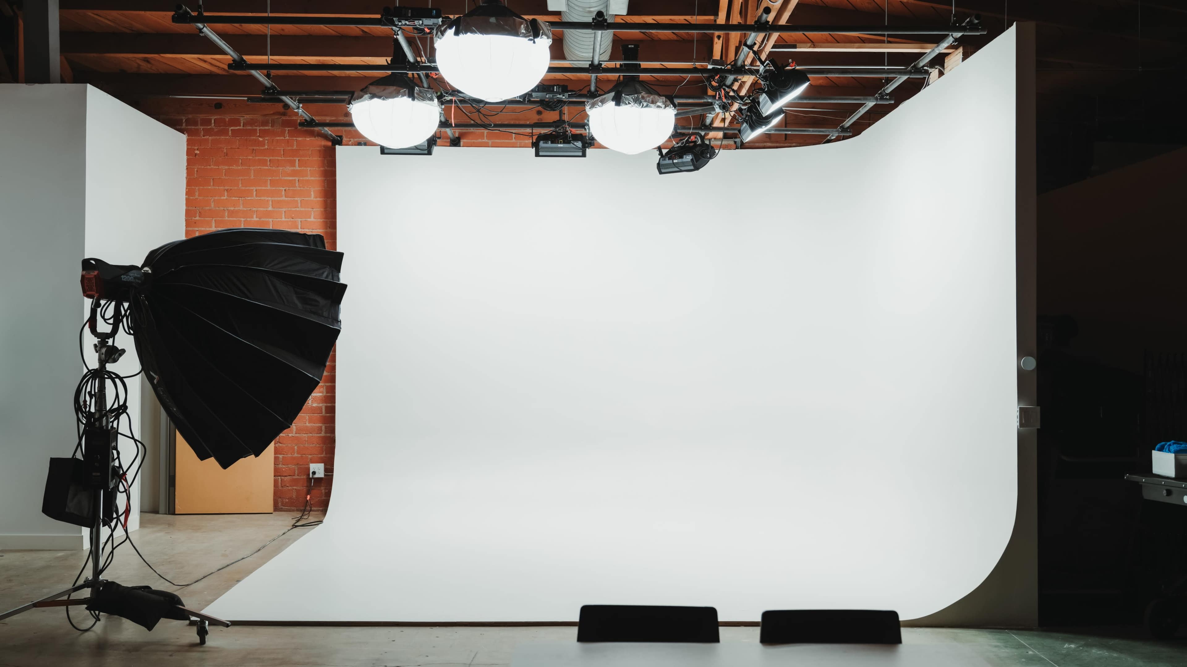 A photography studio features a seamless white backdrop with multiple overhead lights and a large softbox positioned to the side.