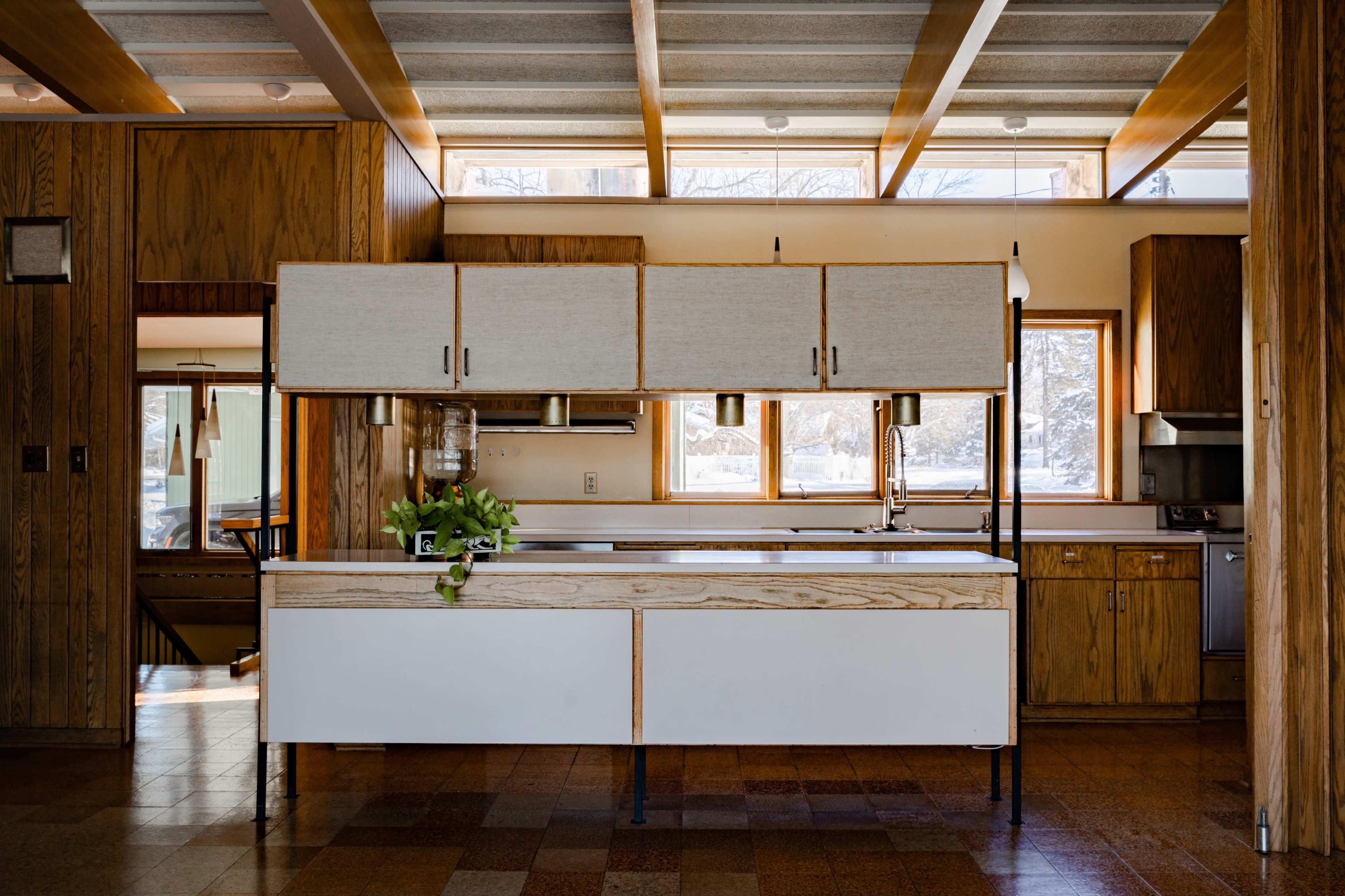 A modern kitchen features a central island with a wooden base, overhead storage cabinets, and large windows allowing natural light to enter.
