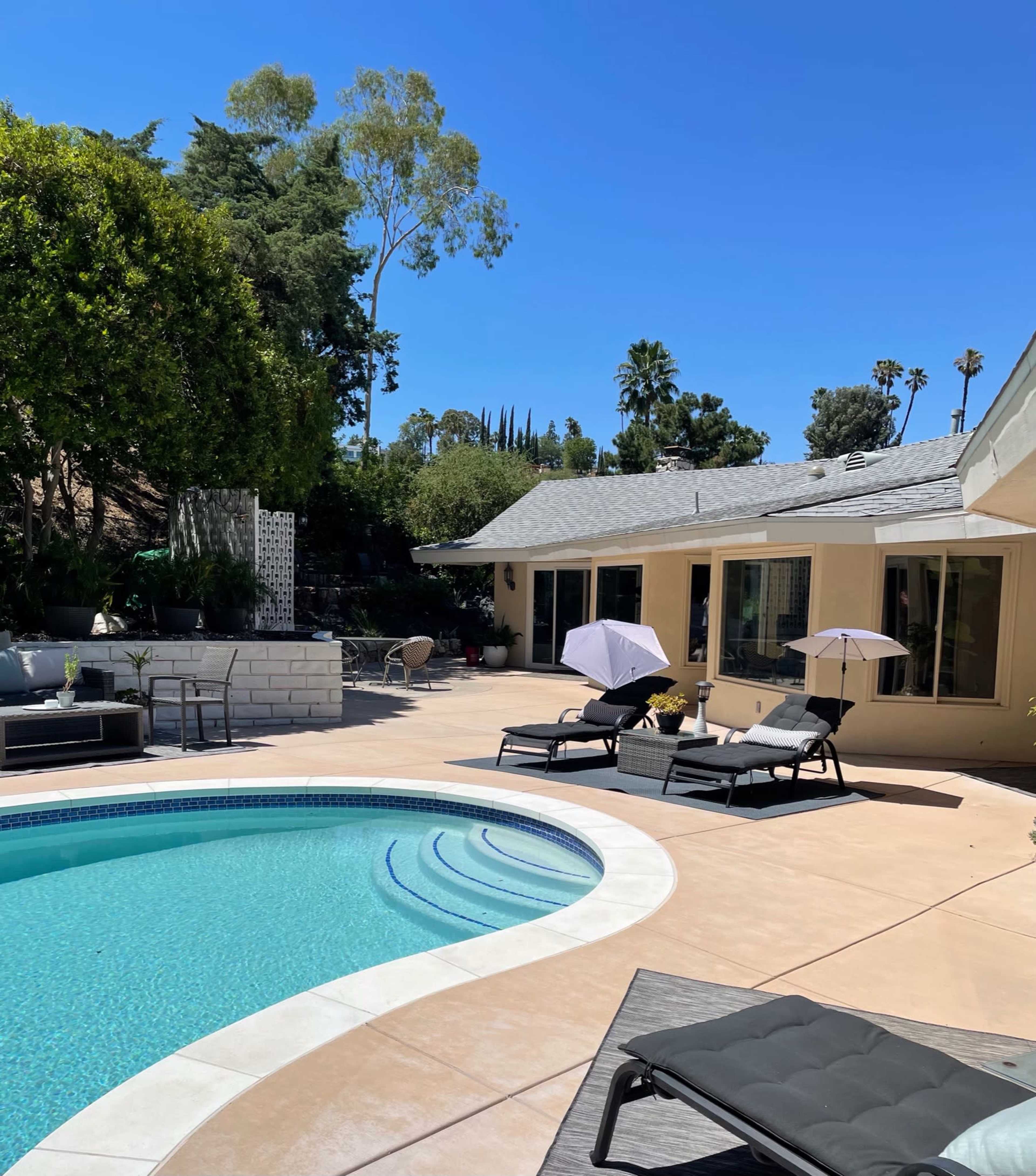 A backyard with a swimming pool, lounge chairs, and a patio area beside a house under clear blue skies.