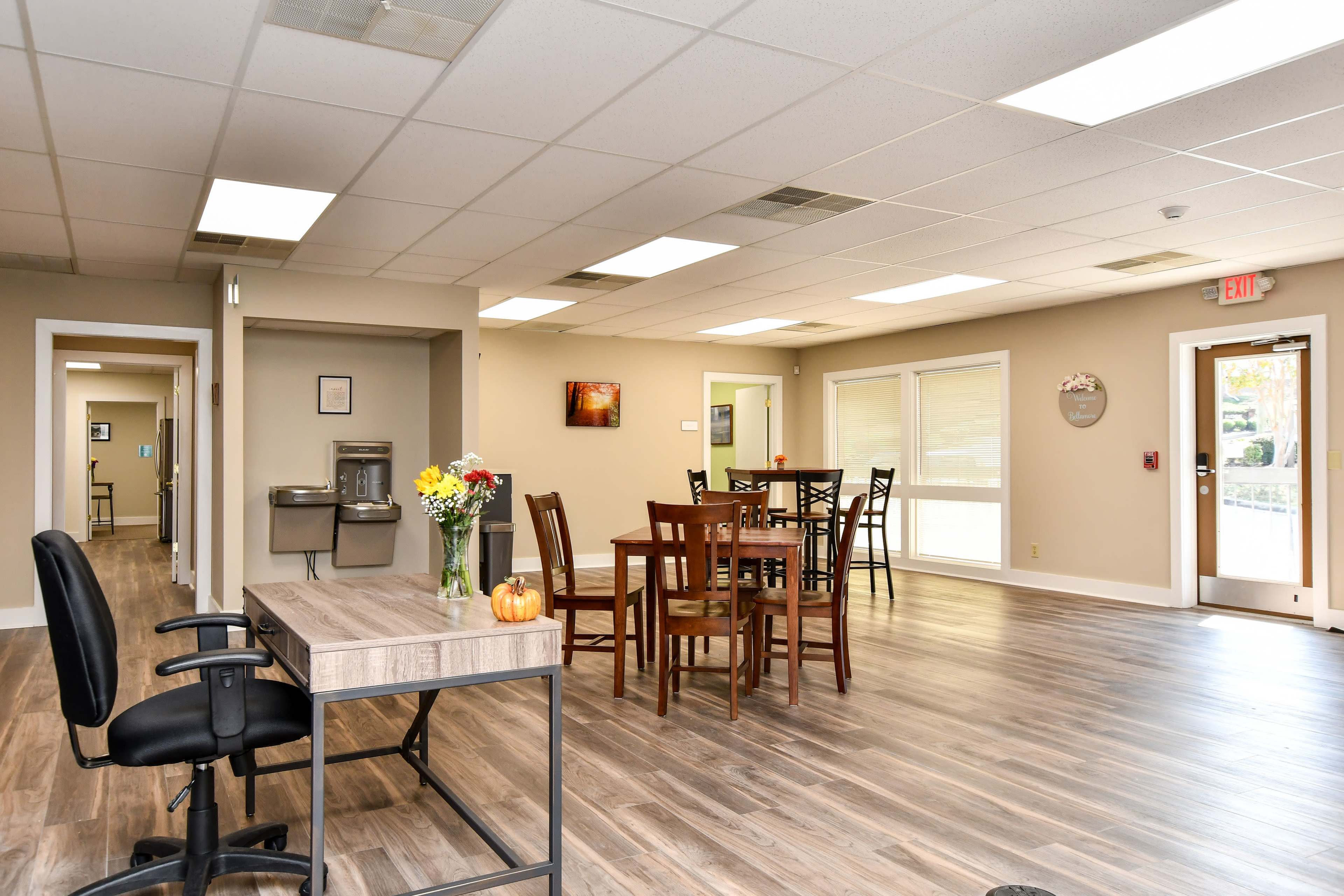 An indoor space featuring tables and chairs, a wooden desk with a swivel chair, a drinking fountain, and large windows allowing natural light.