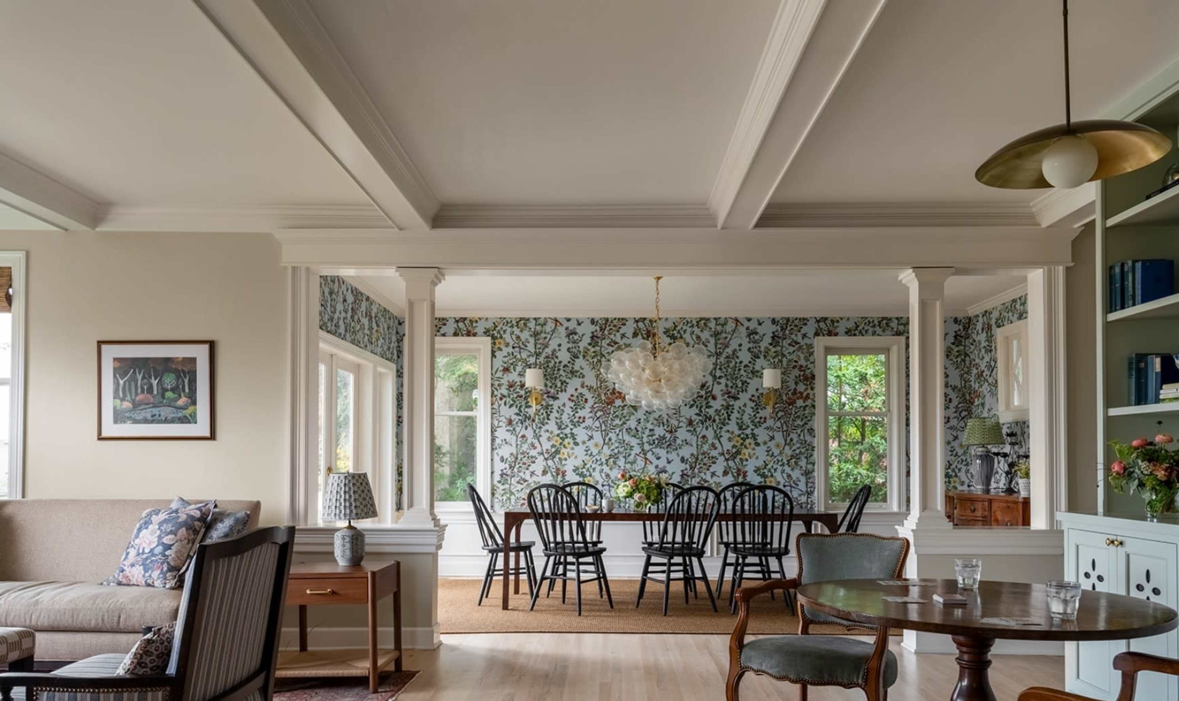 A spacious living area features a dining table set for six, surrounded by floral wallpaper and accented by natural light from large windows.