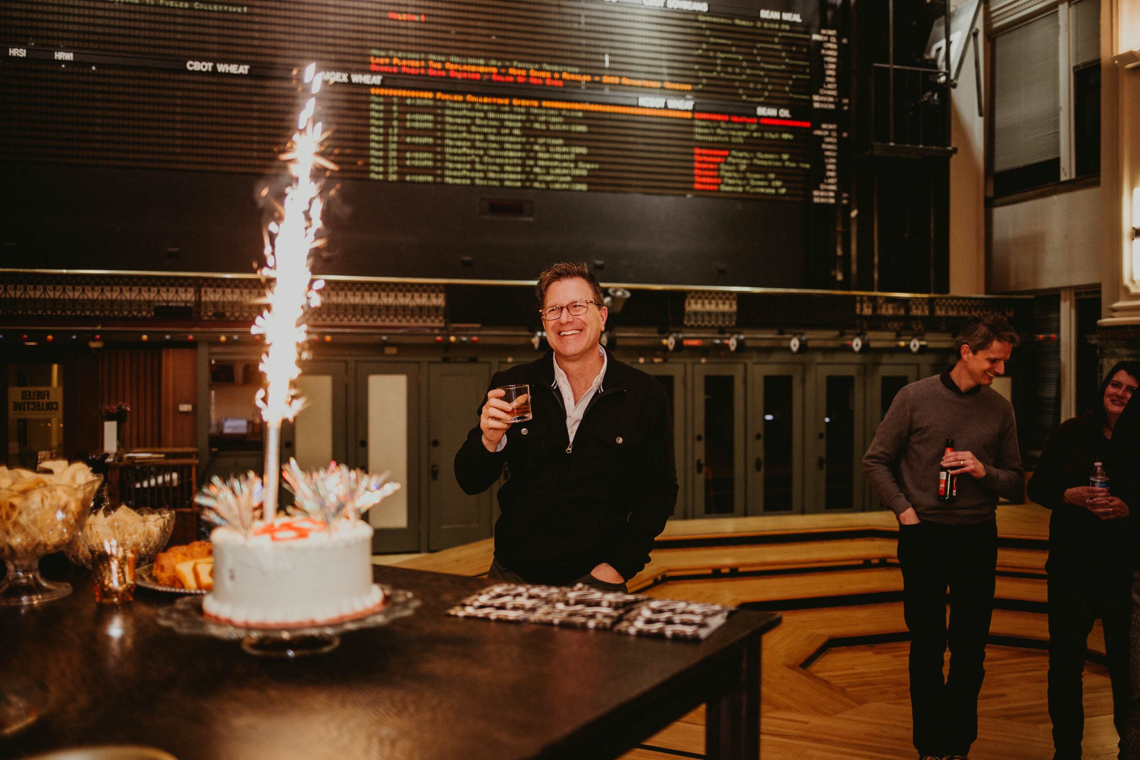 A man stands smiling with a drink in hand next to a cake topped with a sparkler at a celebration in a venue.