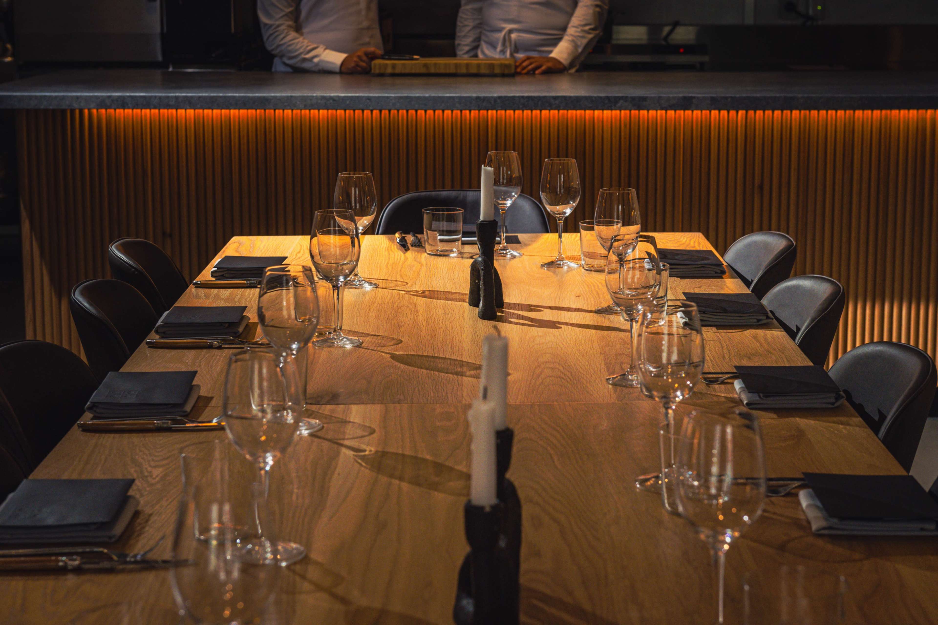 A large wooden dining table is set with glasses and black napkins, illuminated by warm light from the background.