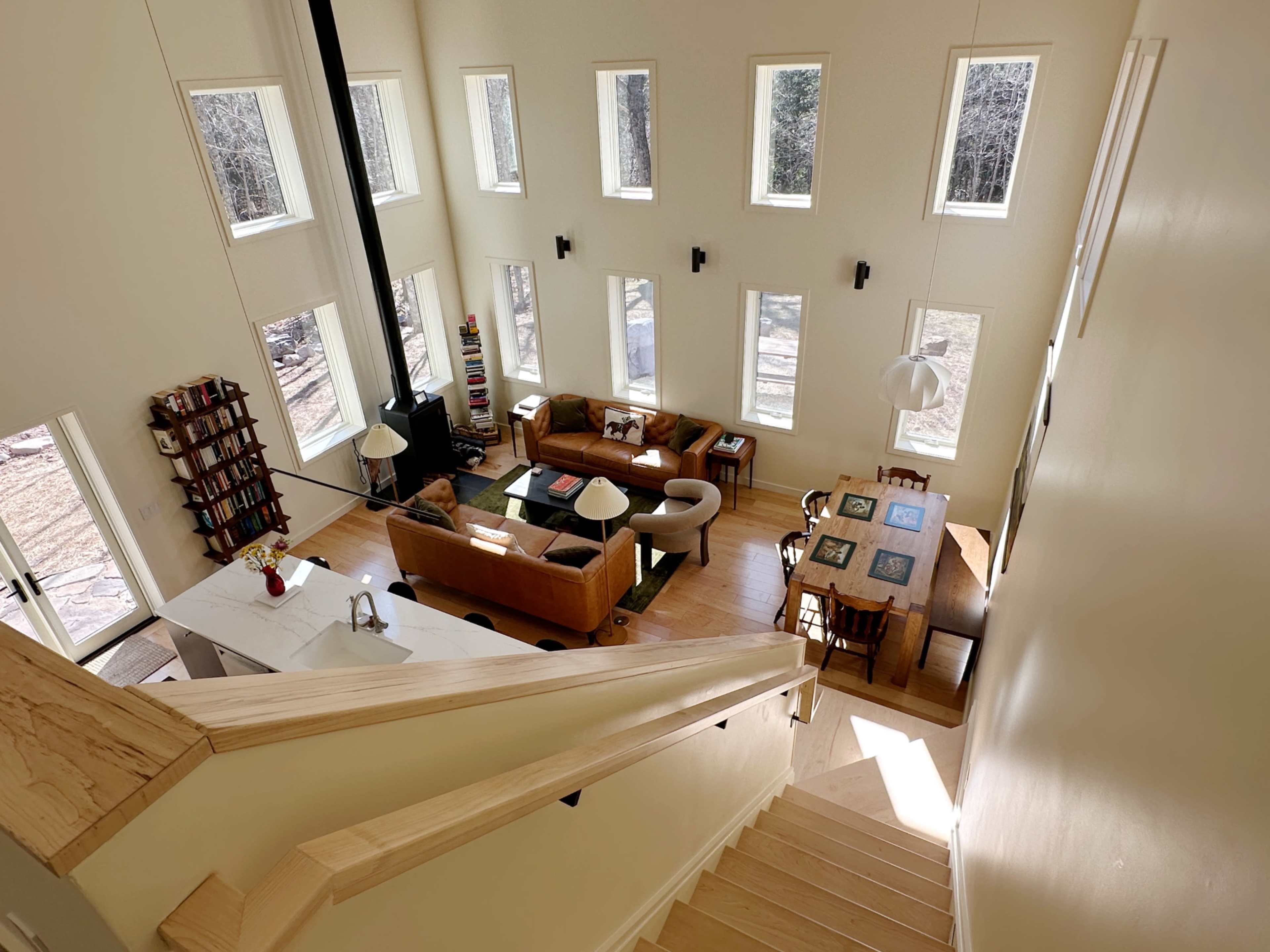 The image shows a spacious living area with large windows, a wooden dining table, and a staircase in a well-lit house.