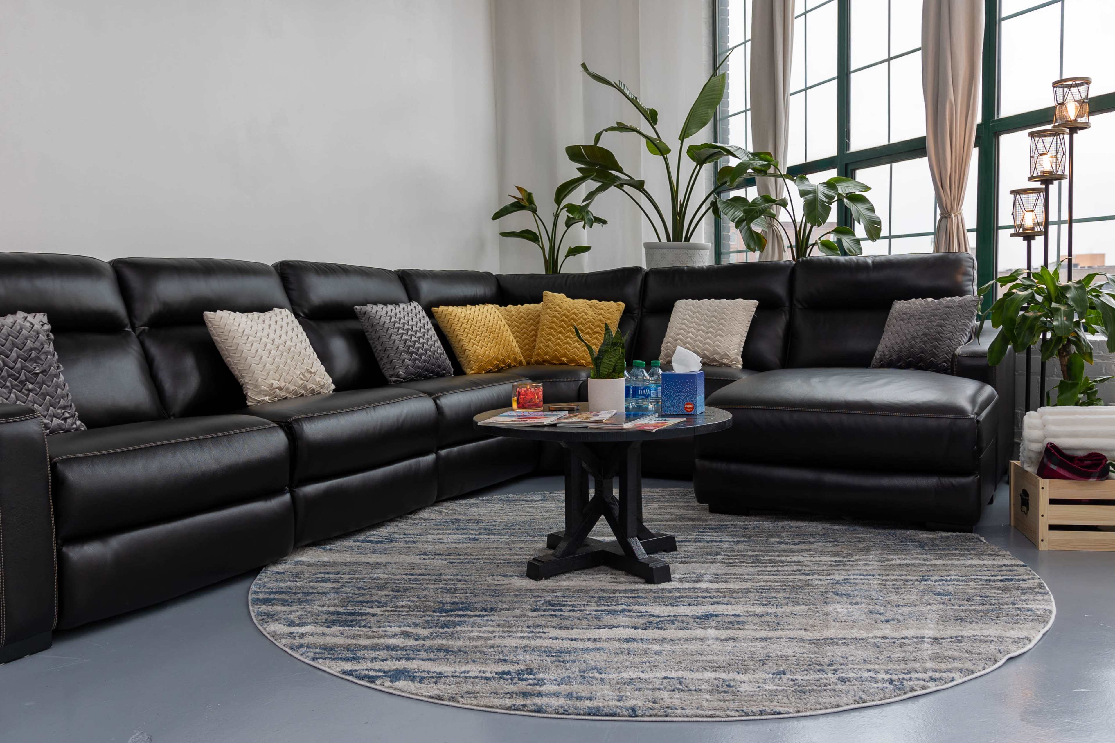 A spacious living area features a large black leather sectional couch, a round area rug, a coffee table with several items on it, and several potted plants by the window.