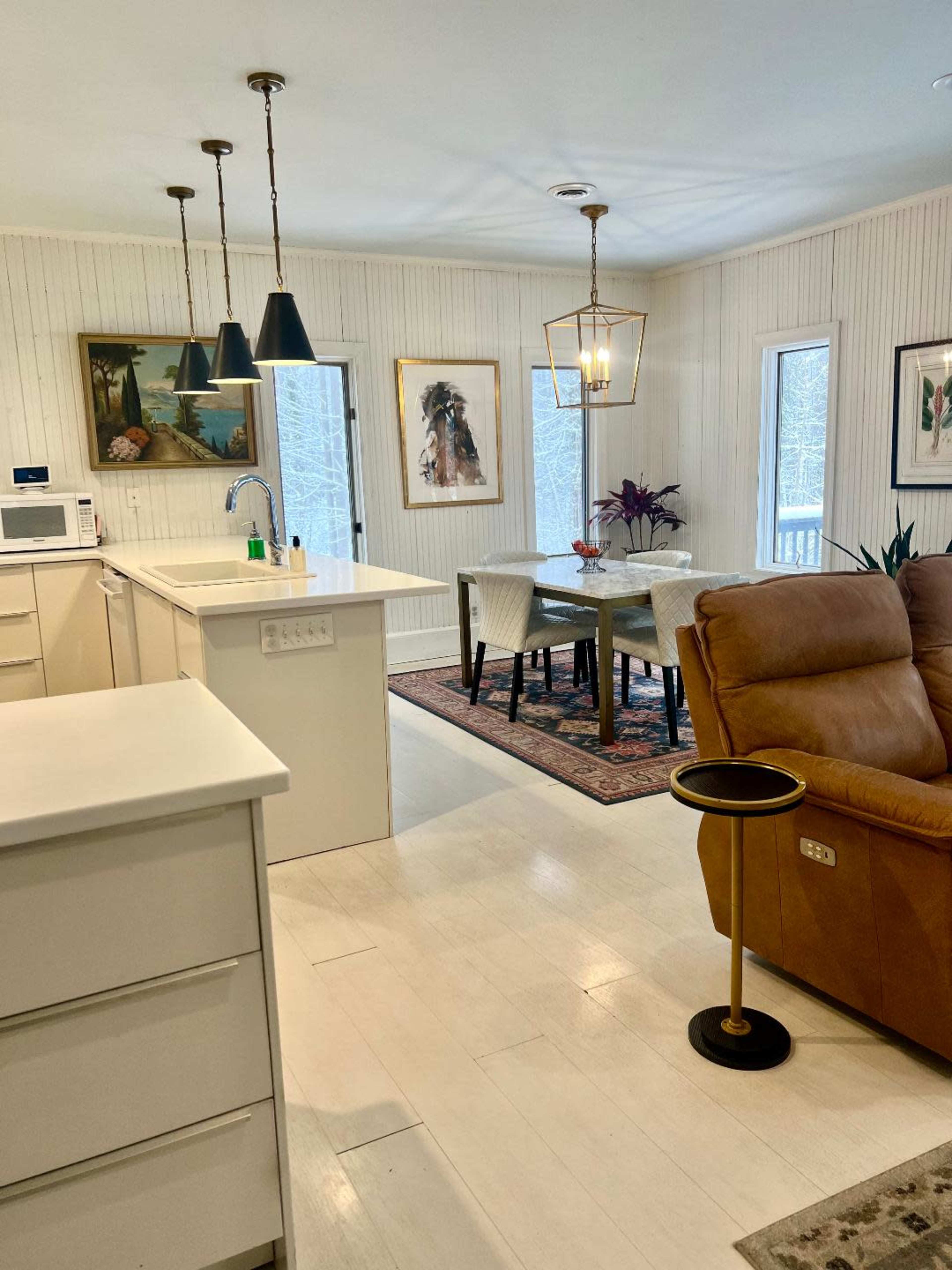 The image shows a bright, modern kitchen and dining area featuring a dining table surrounded by chairs, framed artwork on the walls, and a comfortable recliner in the foreground.