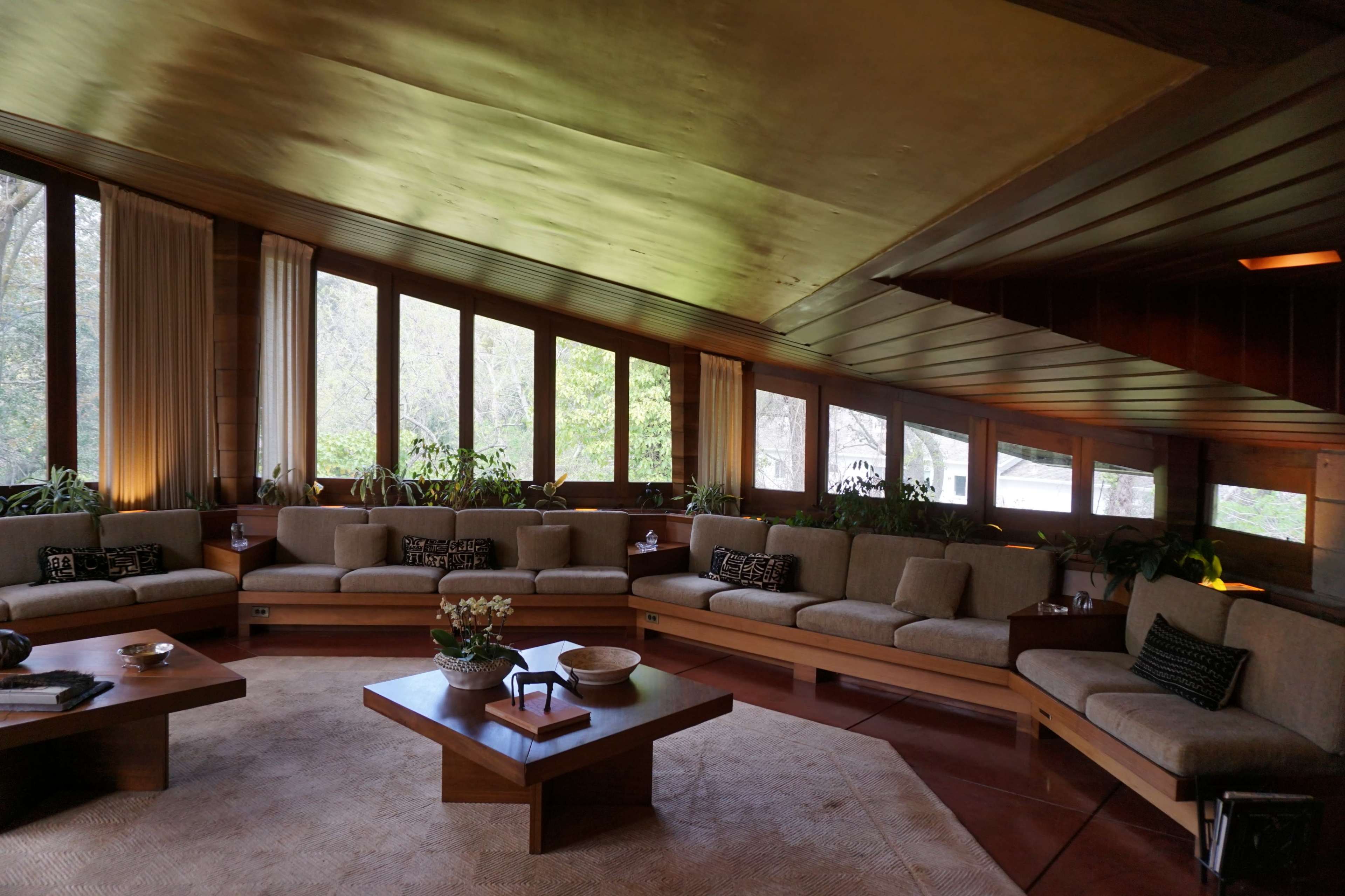 A spacious, modern interior living room with a slanted ceiling, large windows, and a circular seating arrangement around a central coffee table.