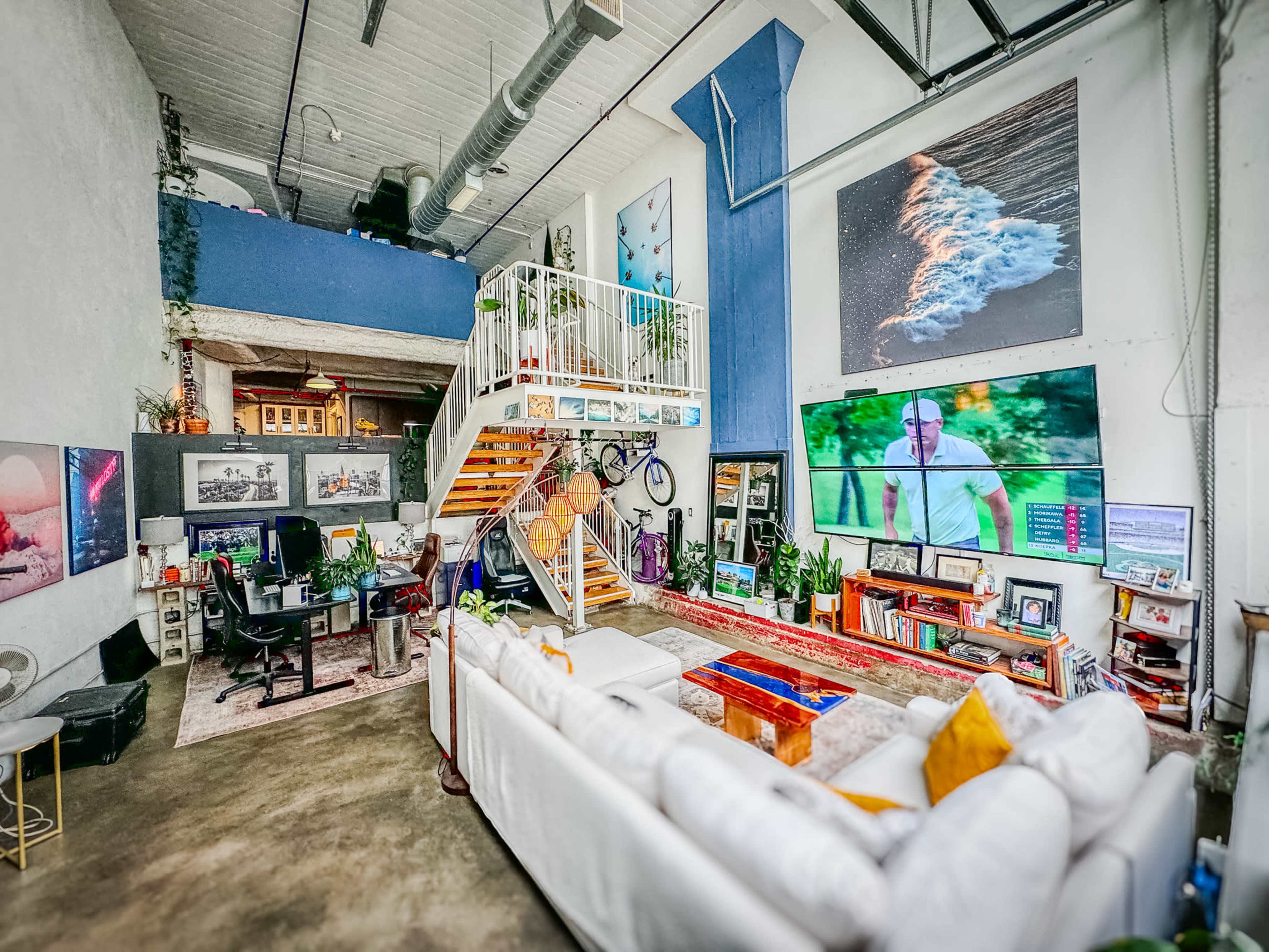 A spacious, modern living area with a staircase leading to a loft, decorated with large artworks and equipped with a television and various plants.