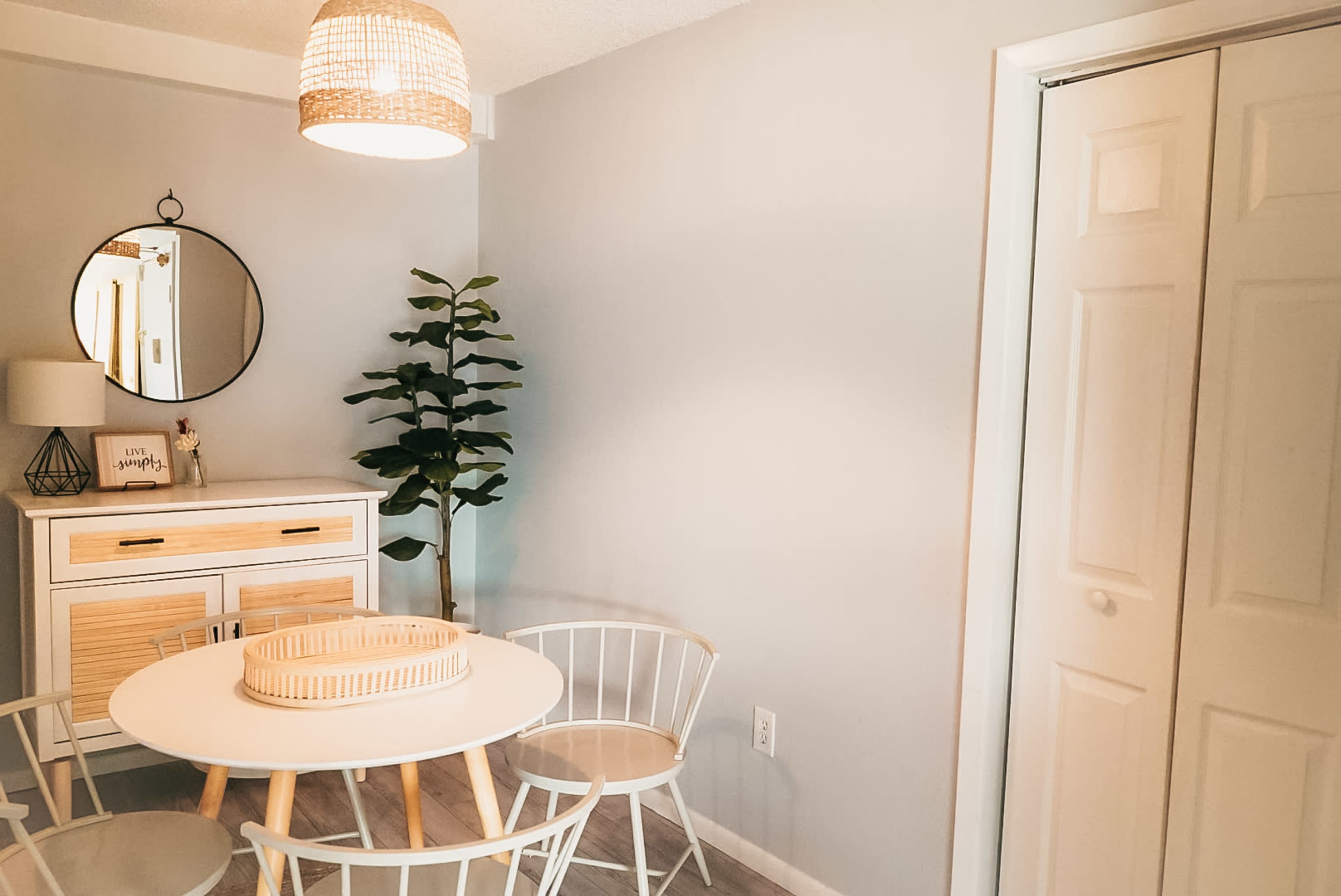 A dining area features a round white table surrounded by four white chairs, a tall green plant in the corner, and a framed mirror above a light-colored sideboard.