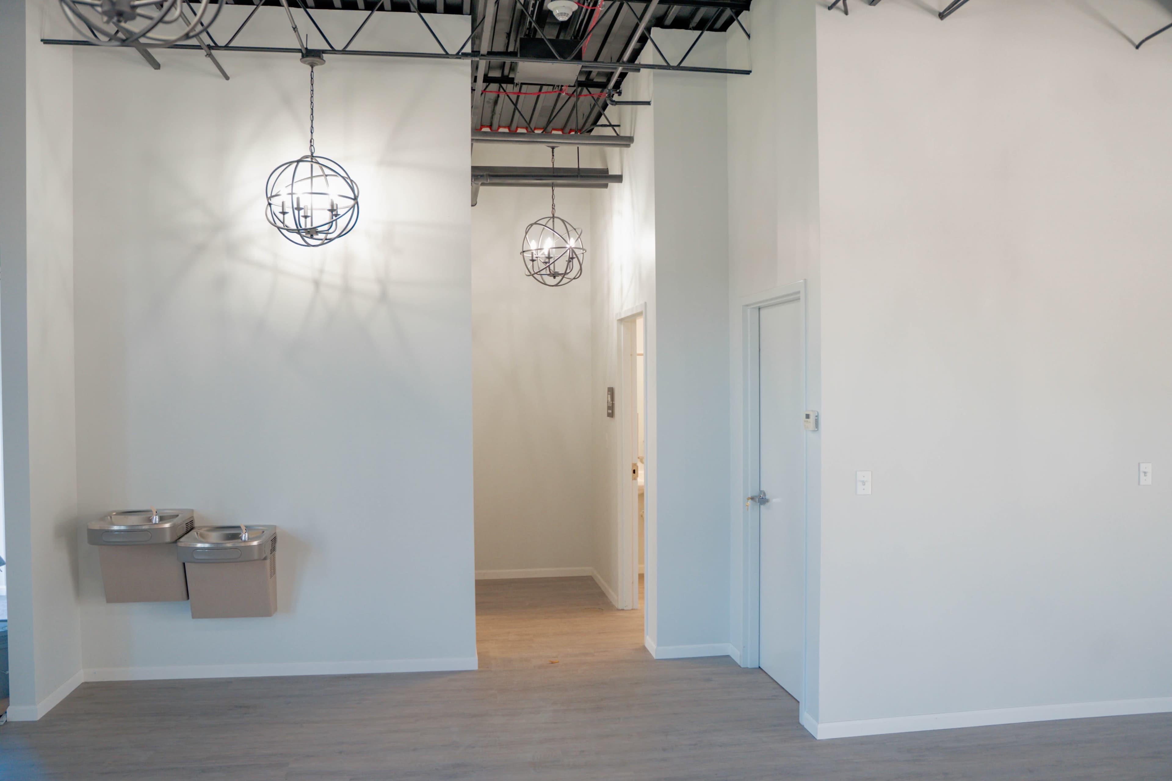 A spacious, well-lit interior with two hanging light fixtures, a hallway leading to a door, and two drinking fountains on the left wall.