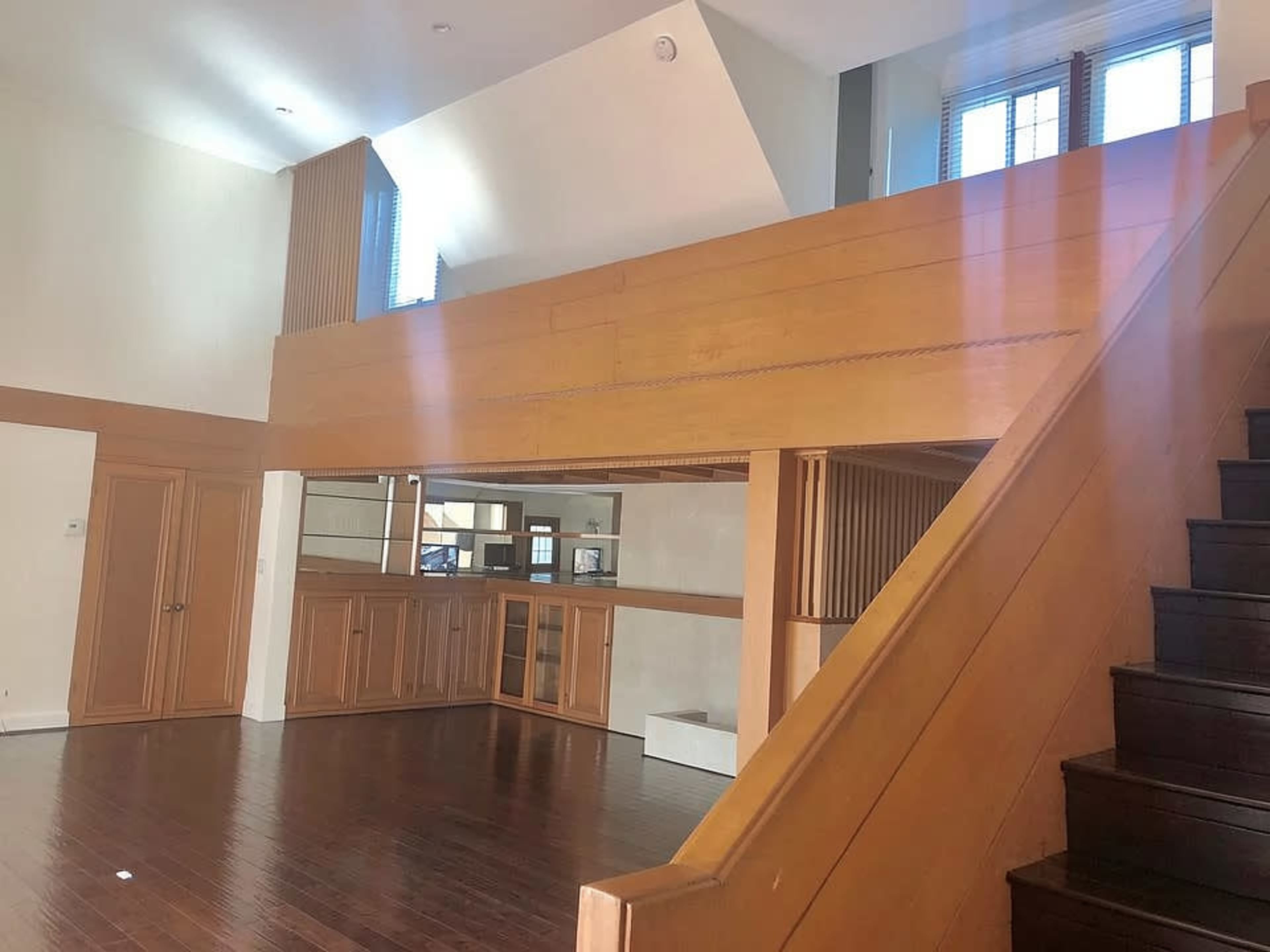The image shows a spacious, two-level interior of a house with wooden features, including a staircase and a balcony overlooking the main floor.