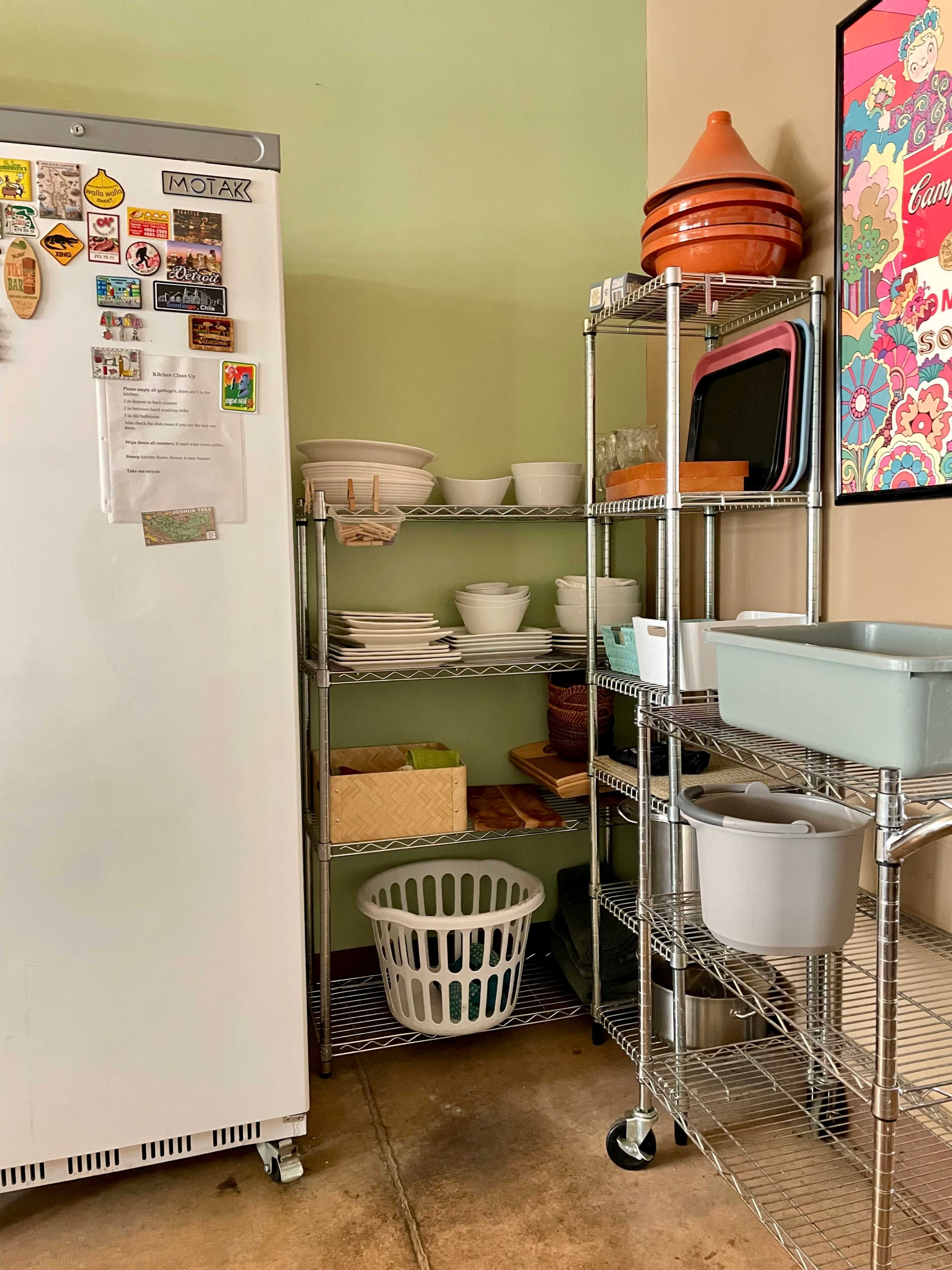 A kitchen corner with a refrigerator, shelves filled with dishes and utensils, and a laundry basket on the floor.
