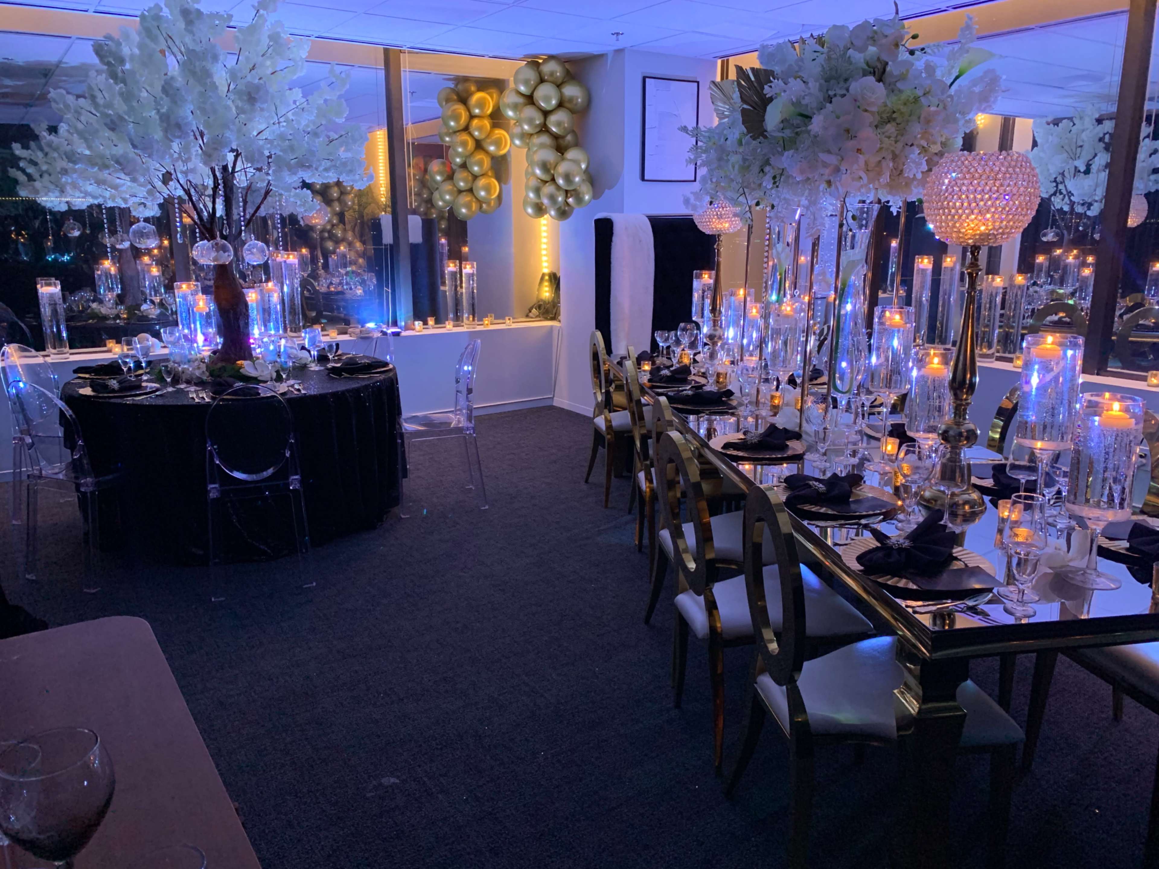 An elegantly decorated dining room with tables set for a formal event, featuring floral centerpieces, candles, and varied lighting.