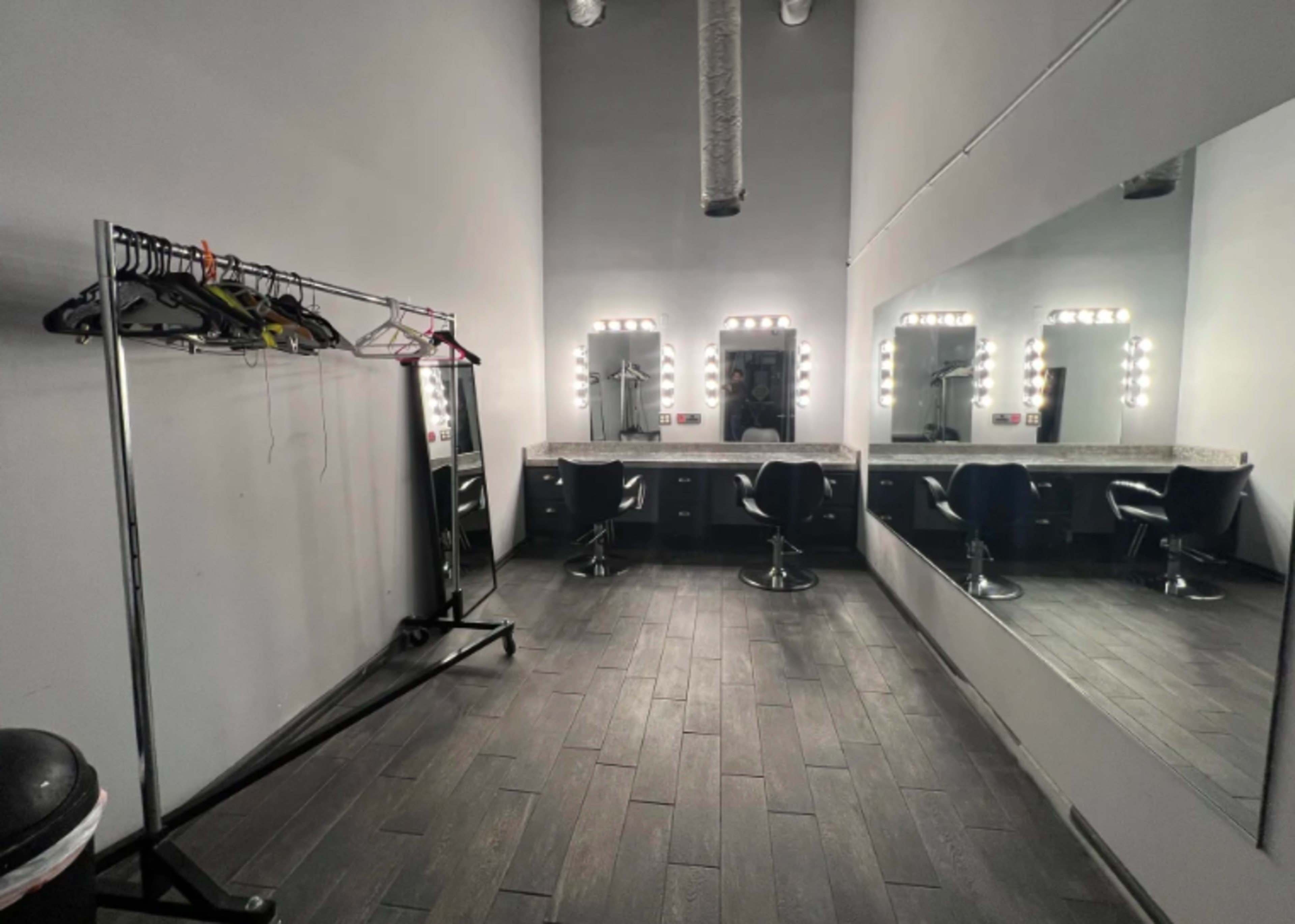 The image shows a dressing room with a long mirror, bright vanity lights, and a clothing rack on the side.