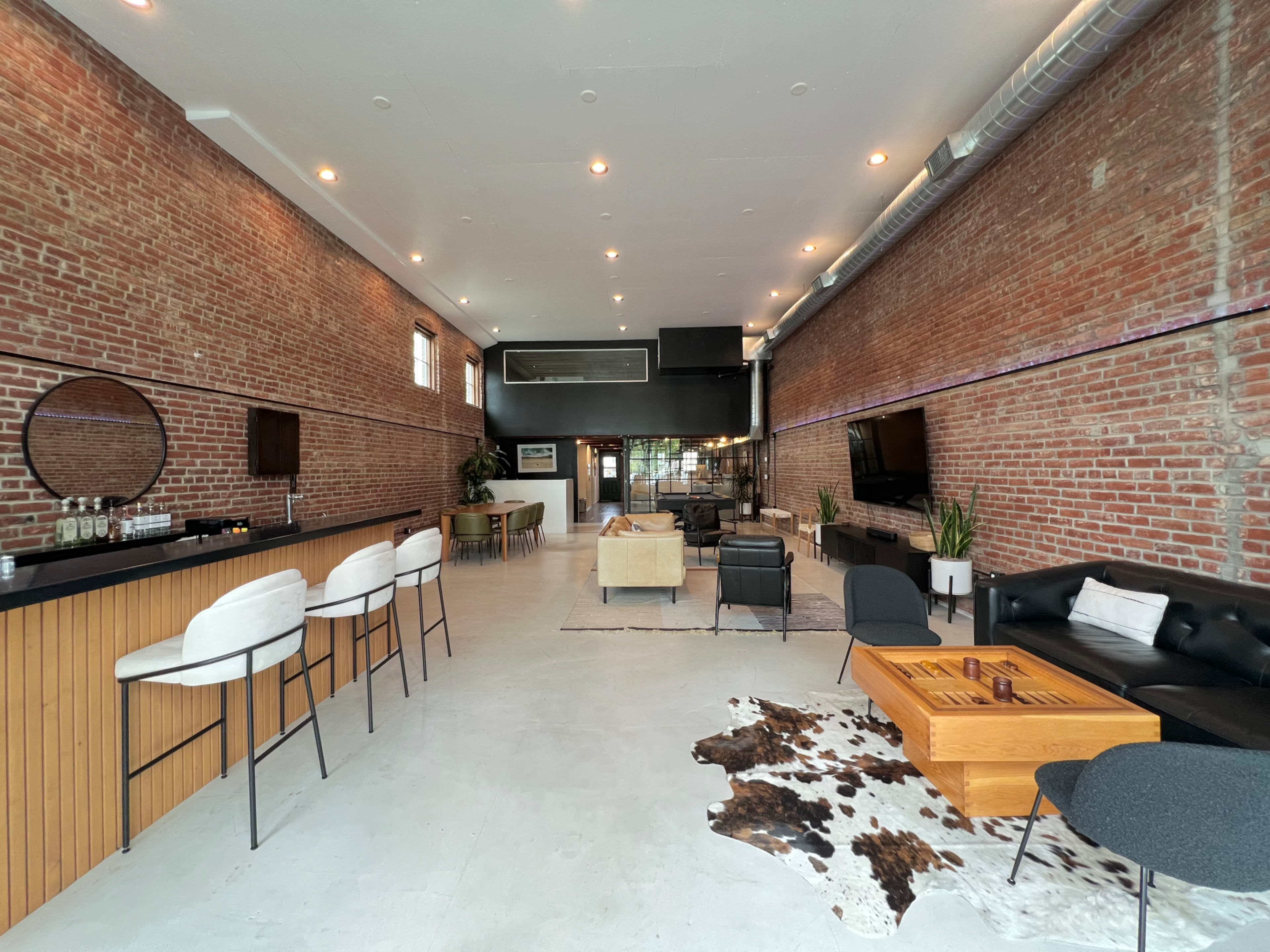 The image shows a spacious interior of a modern lounge or event space with exposed brick walls, a bar area, comfortable seating, and a large window providing natural light.