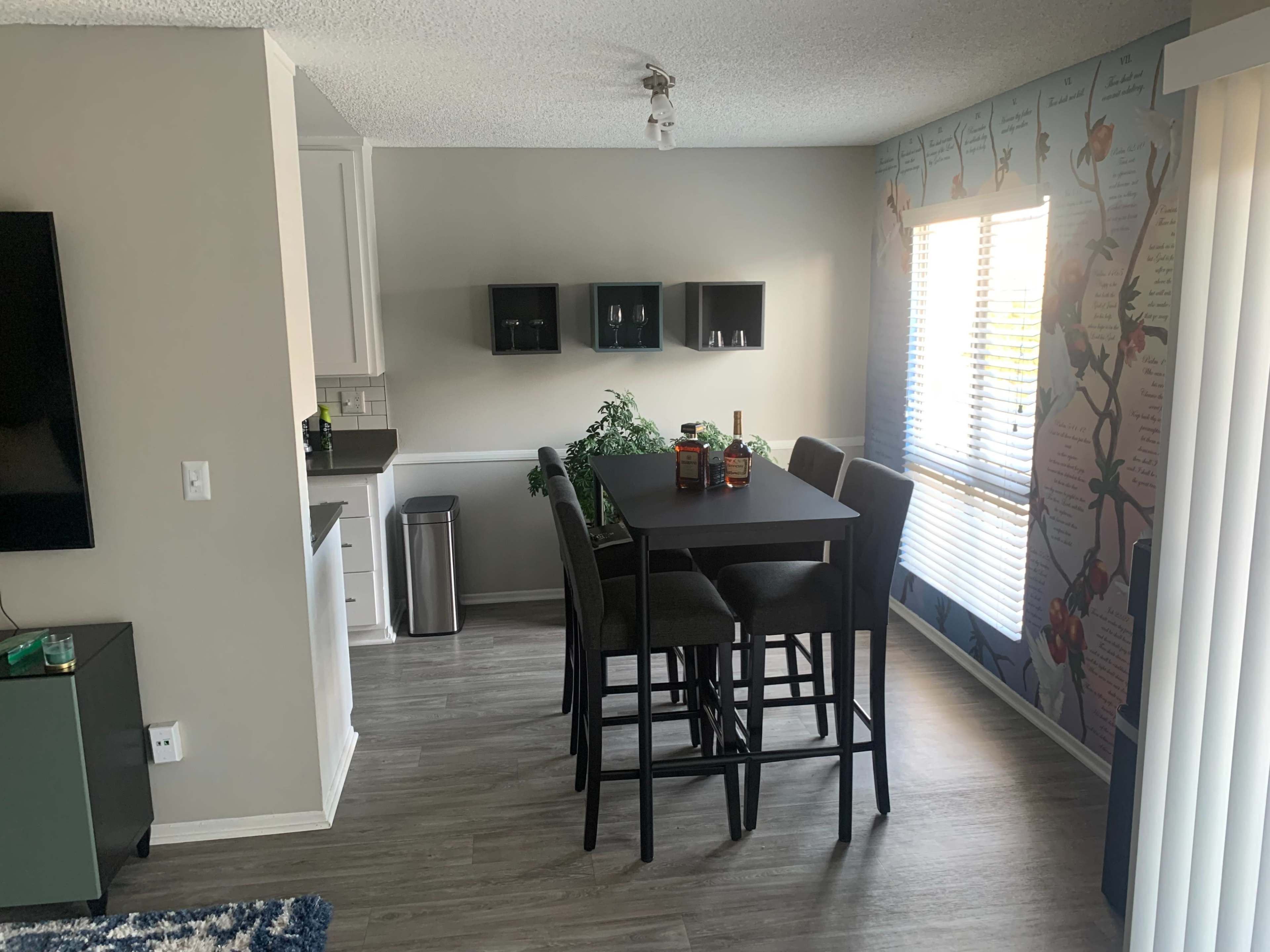 A modern dining area with a tall table and high chairs, adjacent to a kitchen in a well-lit open space featuring a patterned wall.