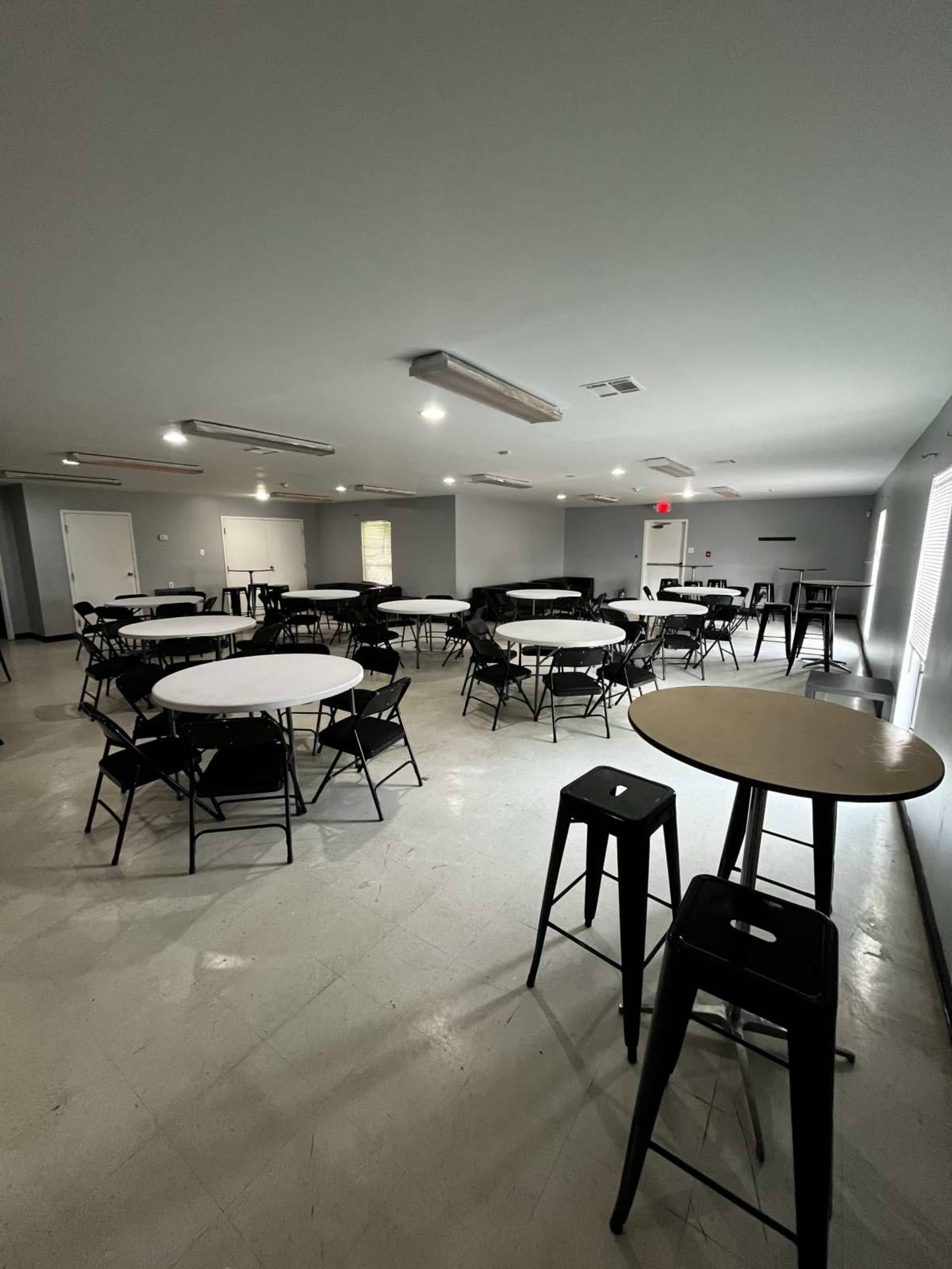 The image shows a spacious room with multiple round tables and black chairs arranged for an event or meeting.