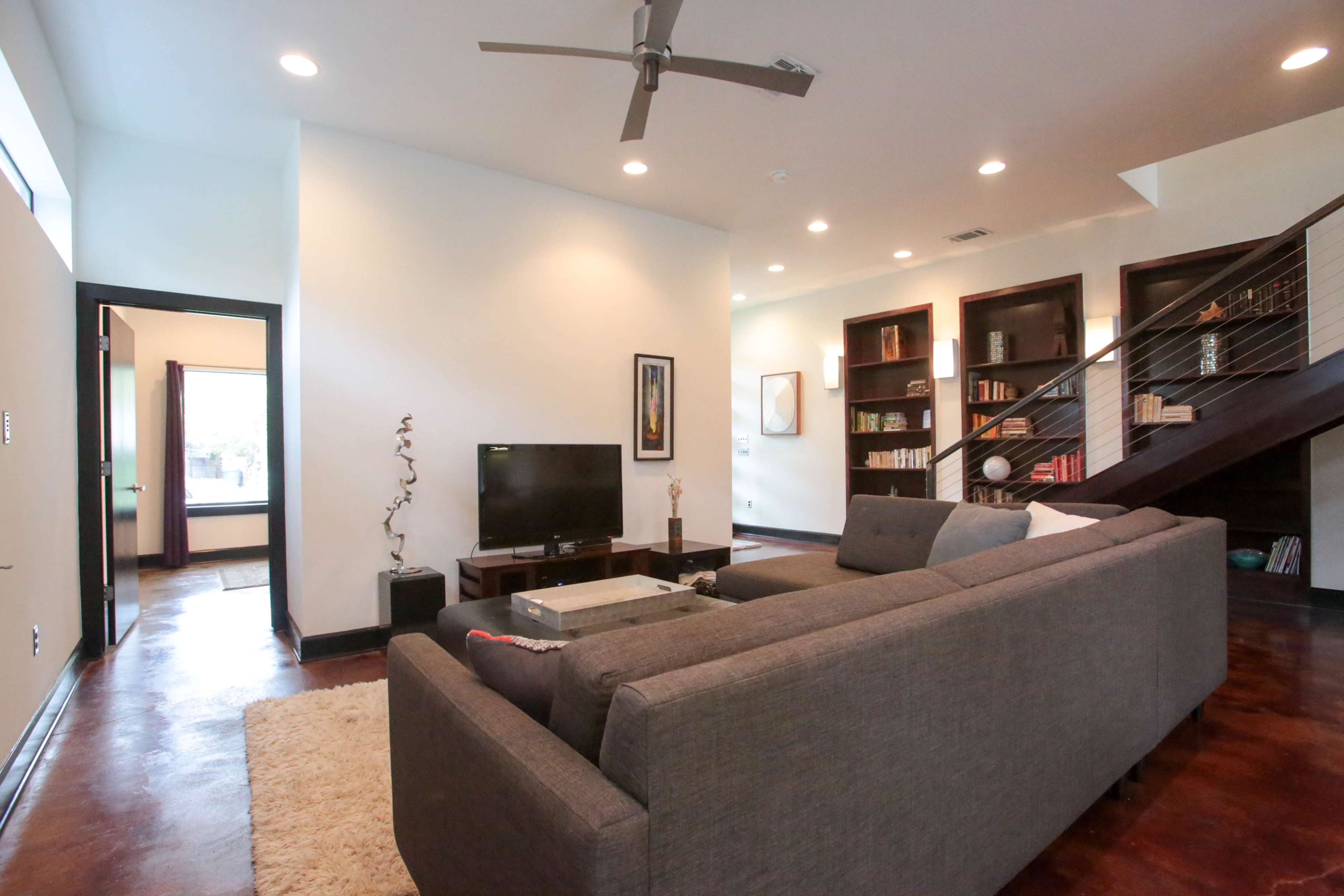 The living room features a sectional sofa, a television on a stand, and shelves filled with books, with a staircase visible in the background.