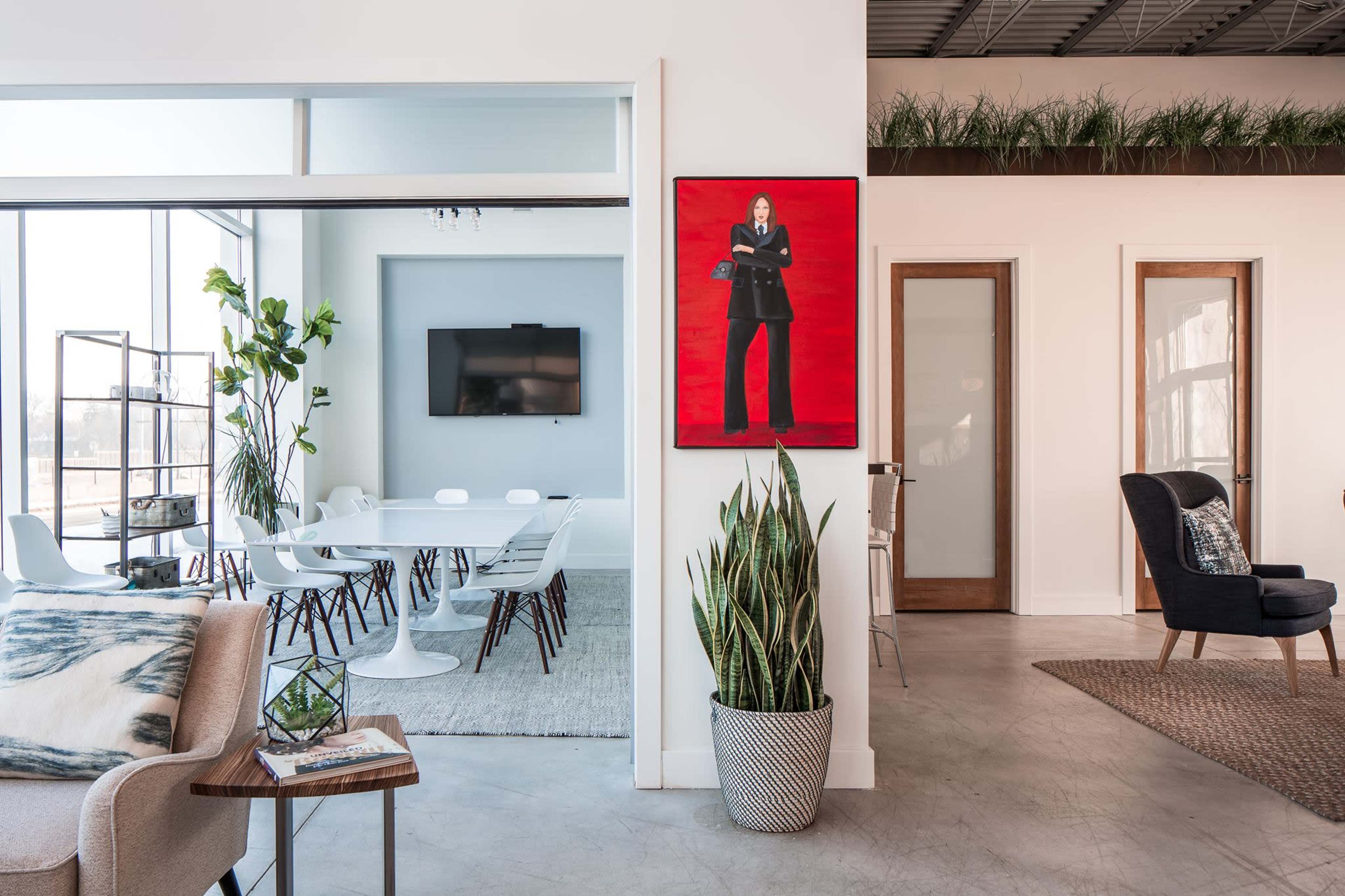 A modern office space features a conference area with a large table, a vibrant red artwork on the wall, and various seating arrangements with plants.