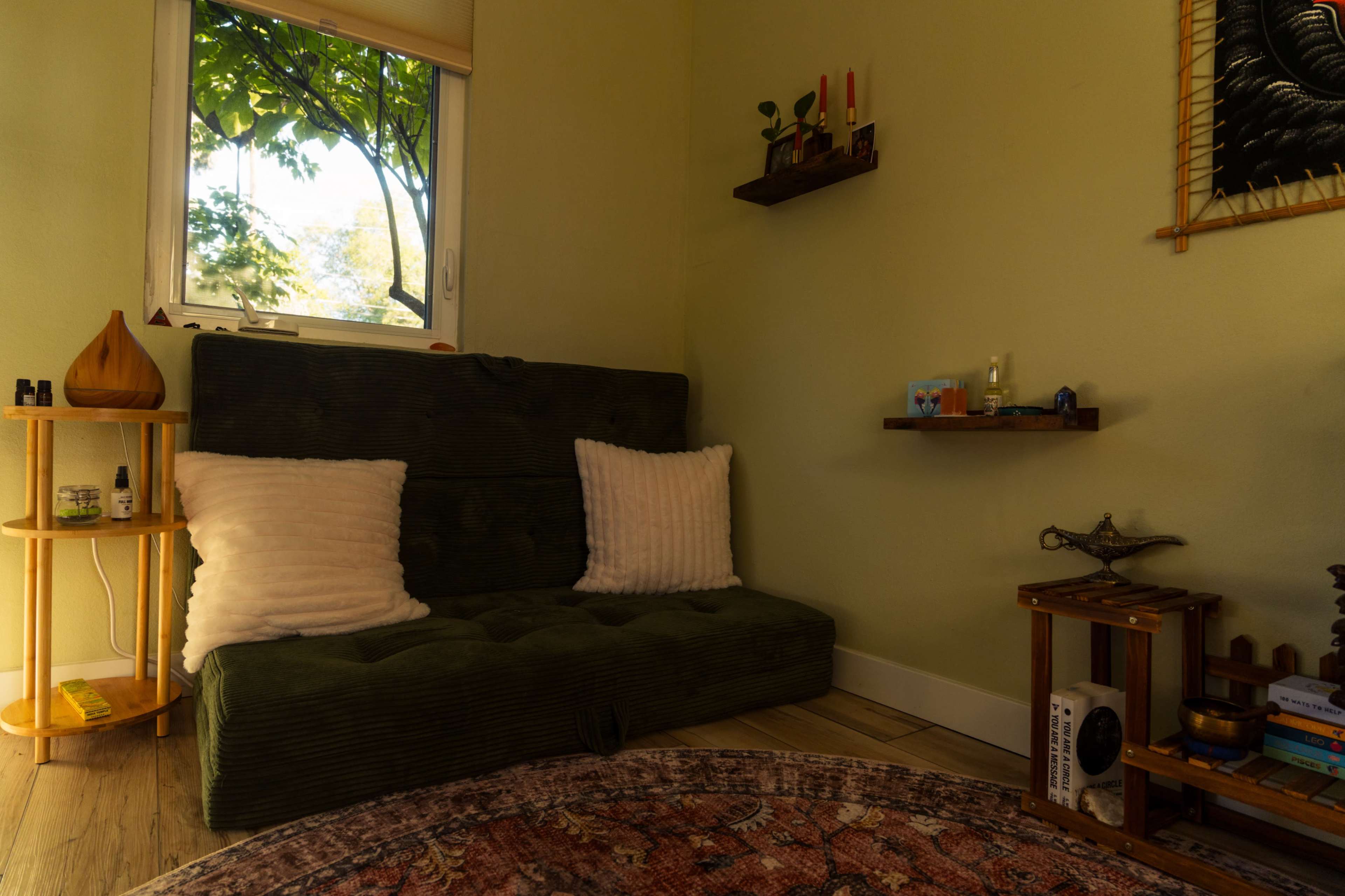 The image shows a cozy corner of a room featuring a green sofa with white pillows, a small shelf with various items, and a window allowing natural light to illuminate the space.