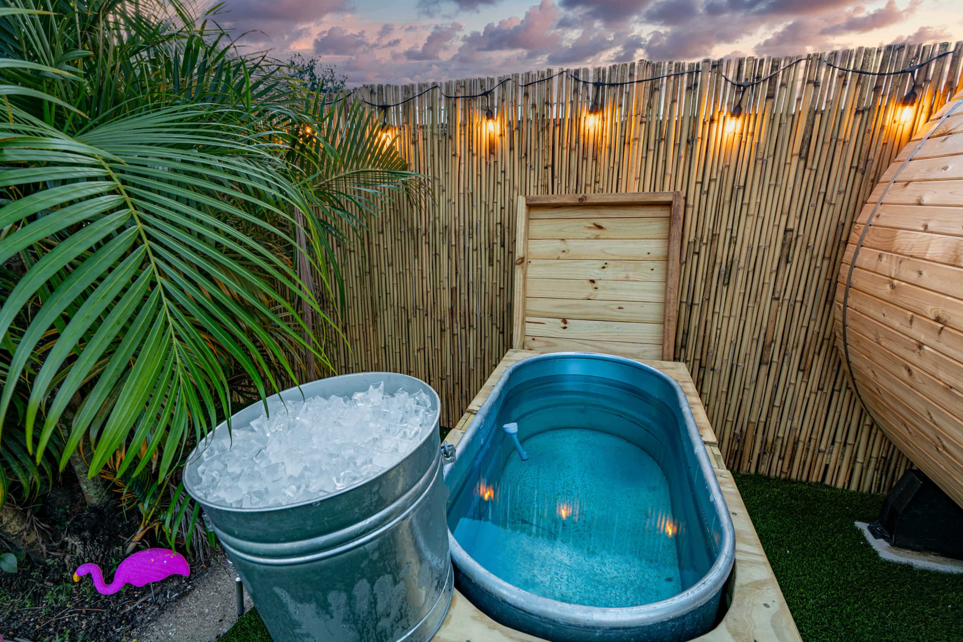 The image shows a wooden hot tub surrounded by a bamboo fence, with a metal bucket filled with ice nearby and a pink flamingo decoration in the corner.