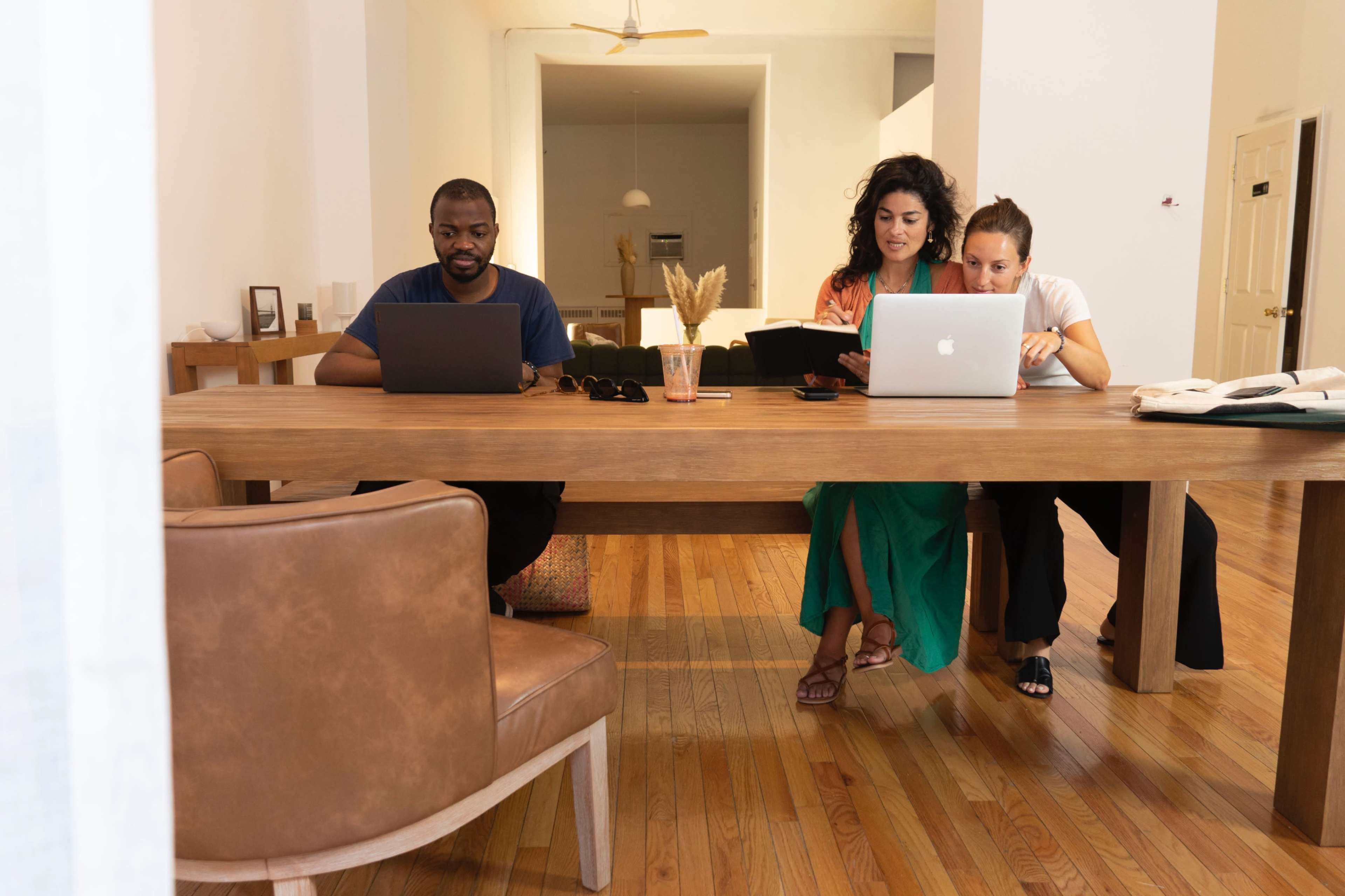 Three individuals are seated at a wooden table, each using a laptop in a bright, spacious room.