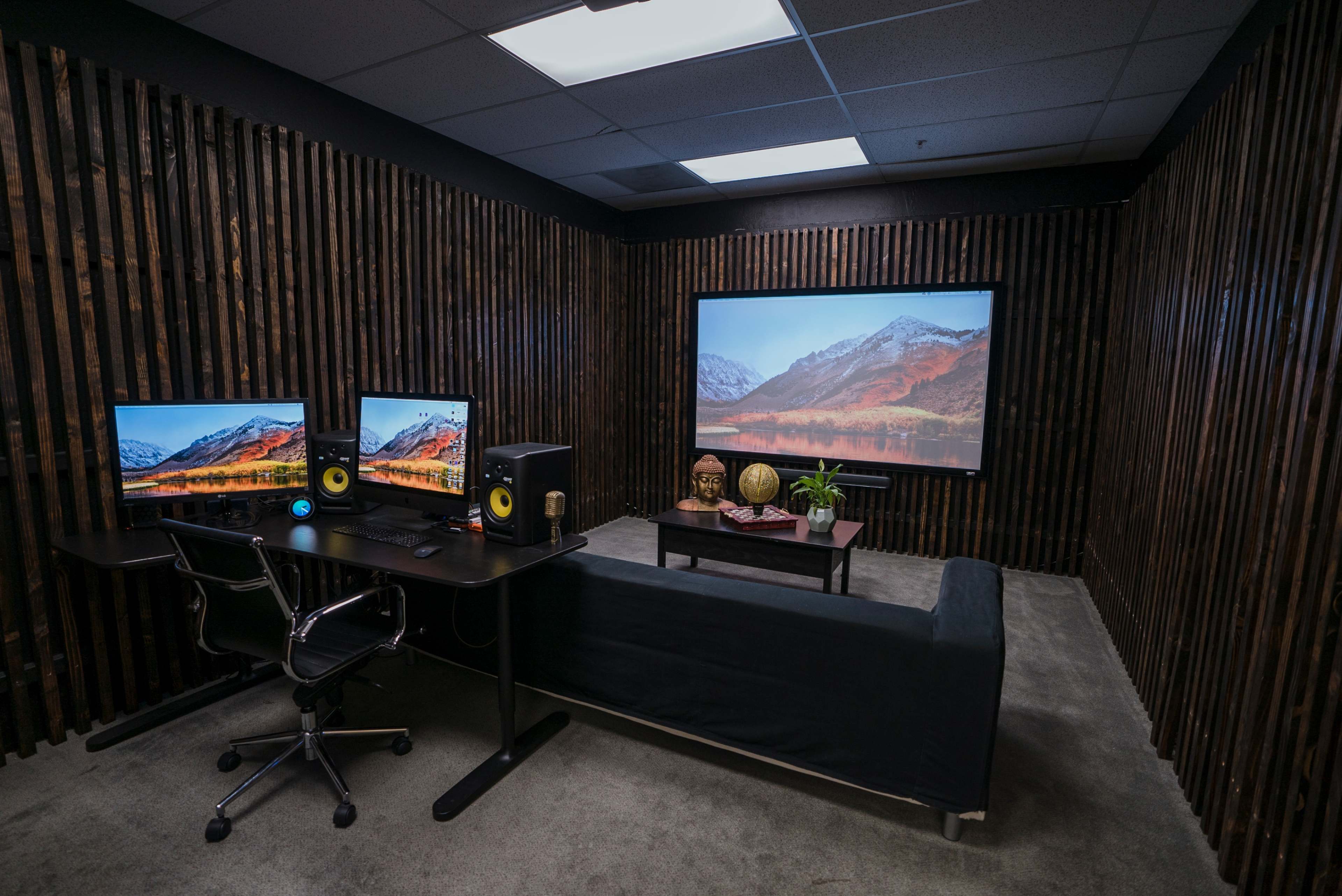 A modern office space features two computer monitors on a desk, a black couch, and a large screen with a nature scene, all set against wooden paneling.
