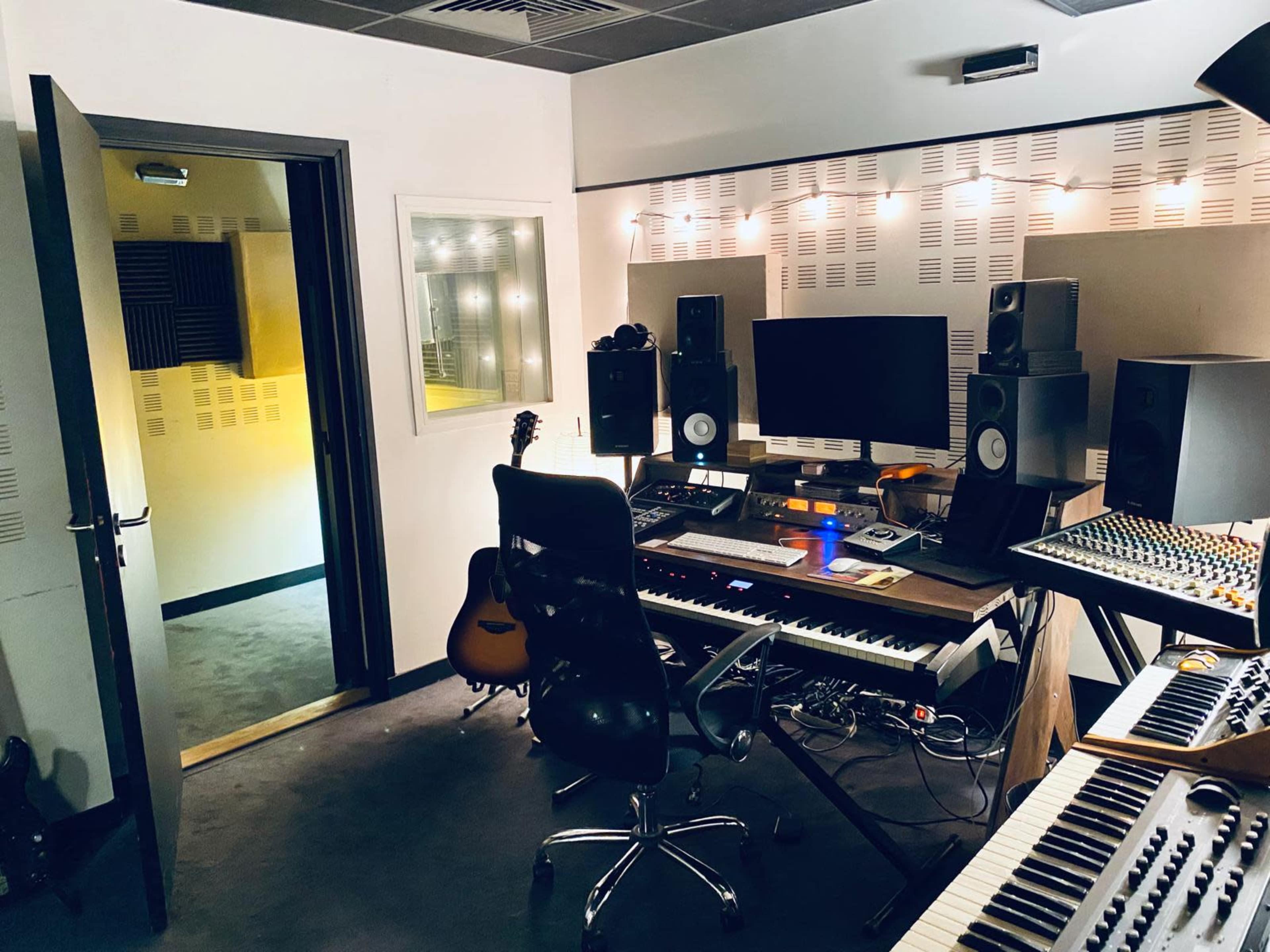 The image shows a recording studio featuring a desk with musical instruments, a computer, and various audio equipment, with soundproofing materials on the walls.