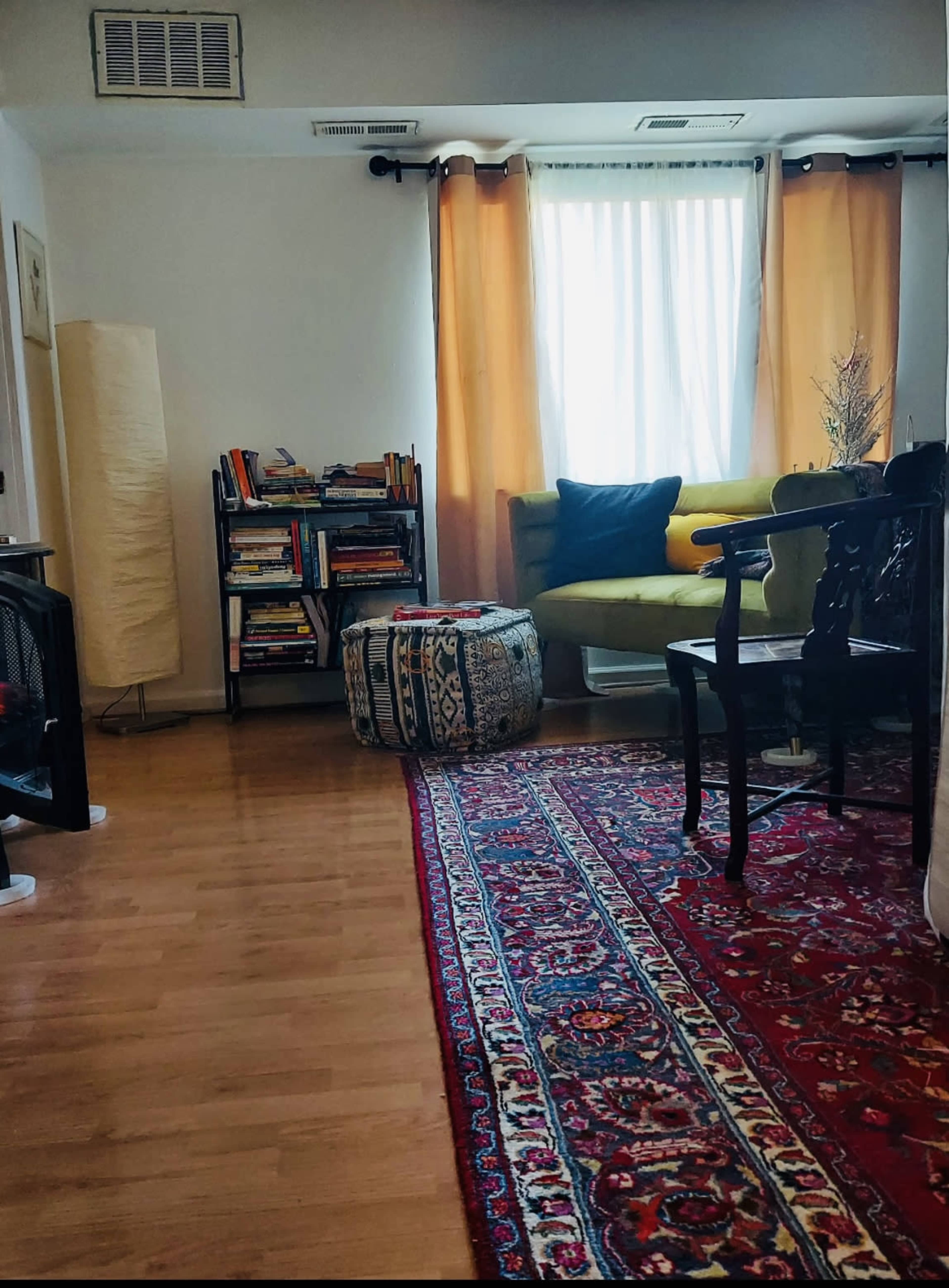 A well-lit living room features a green sofa, a bookshelf filled with books, a patterned ottoman, and a decorative rug on the wooden floor.