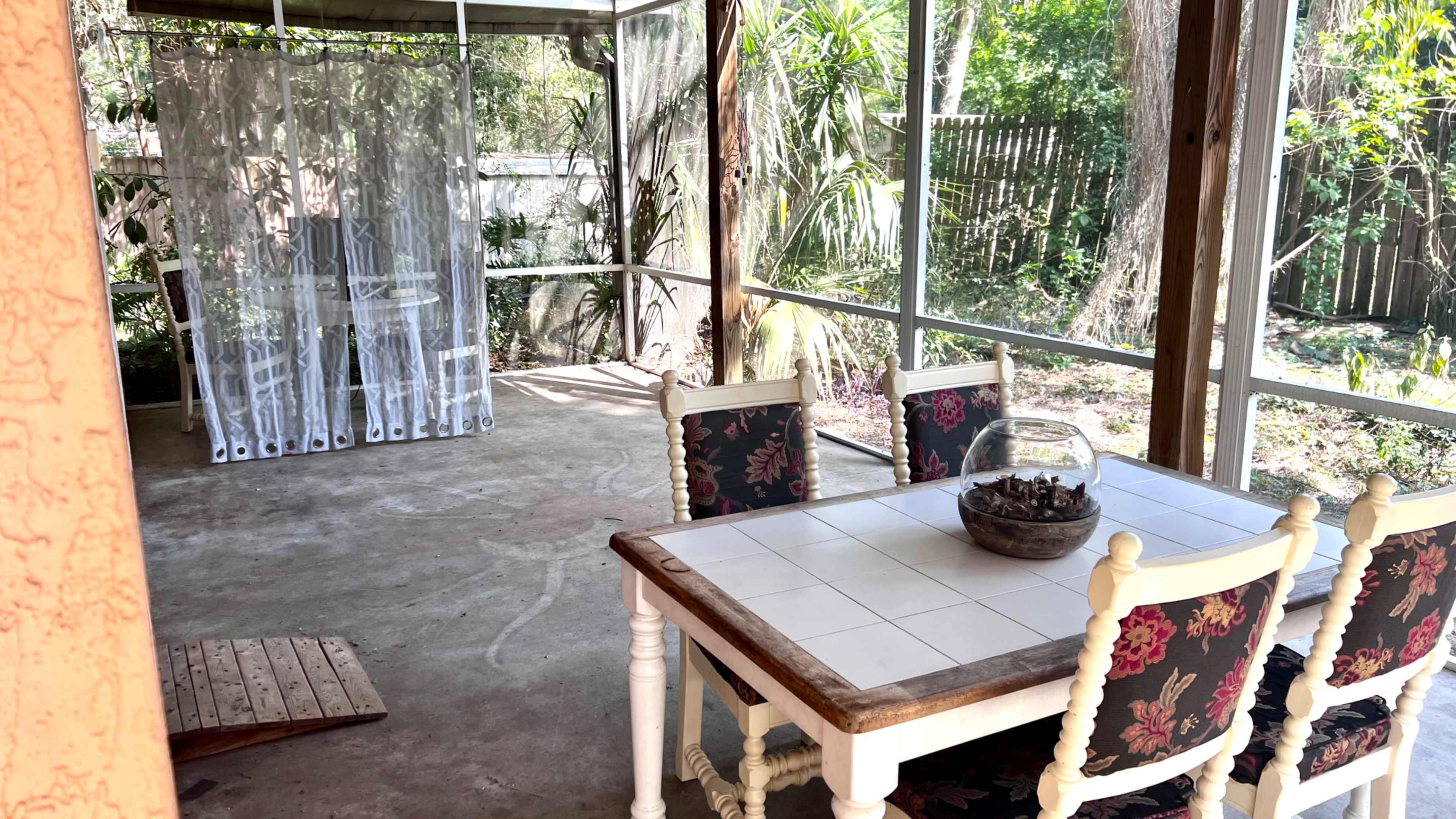 A screened-in porch features a table with four chairs and a decorative bowl on top, surrounded by greenery and a sheer curtain.