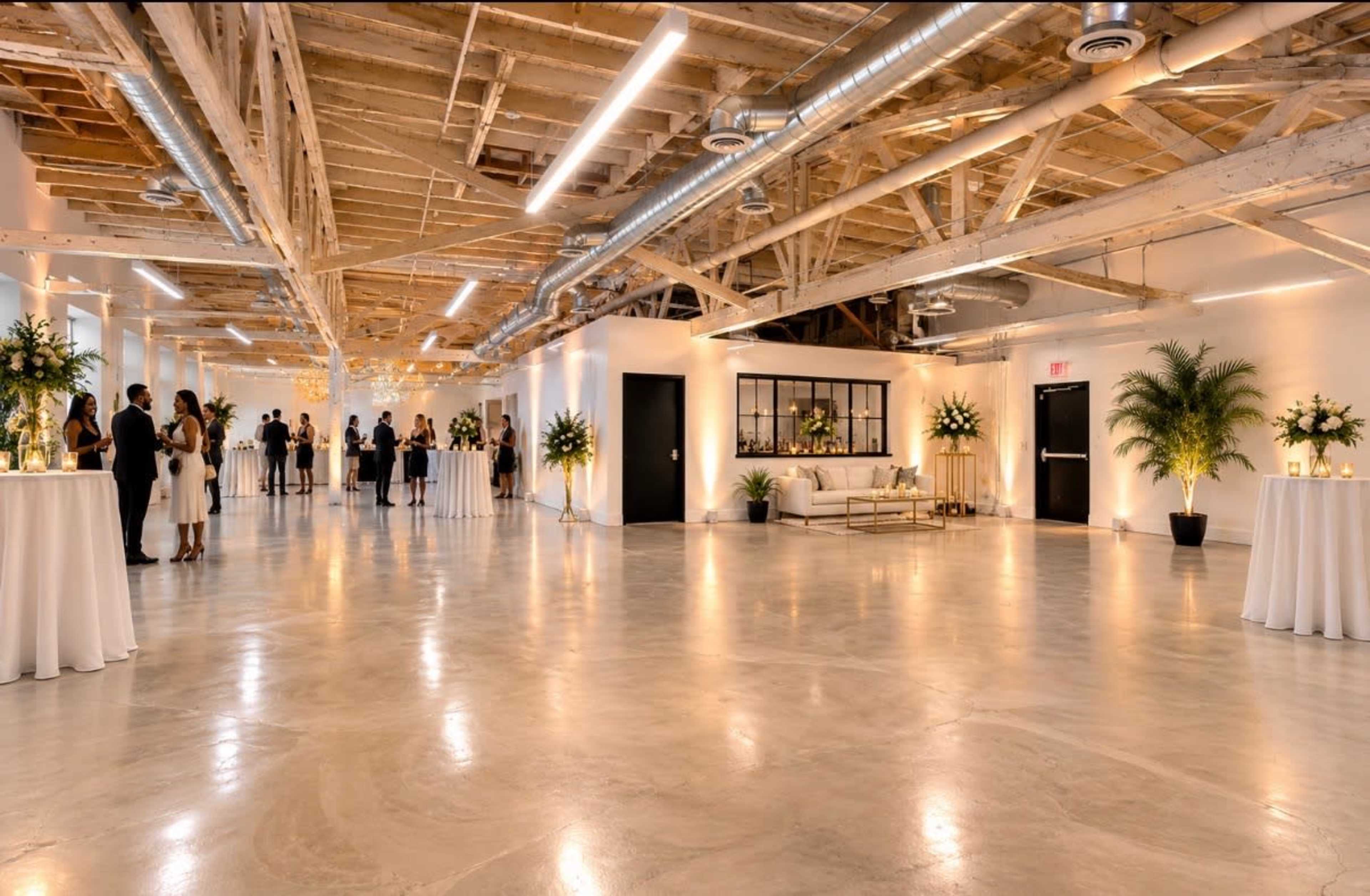 The image shows a spacious, elegantly designed event venue with high ceilings, exposed wooden beams, and guests mingling around cocktail tables.