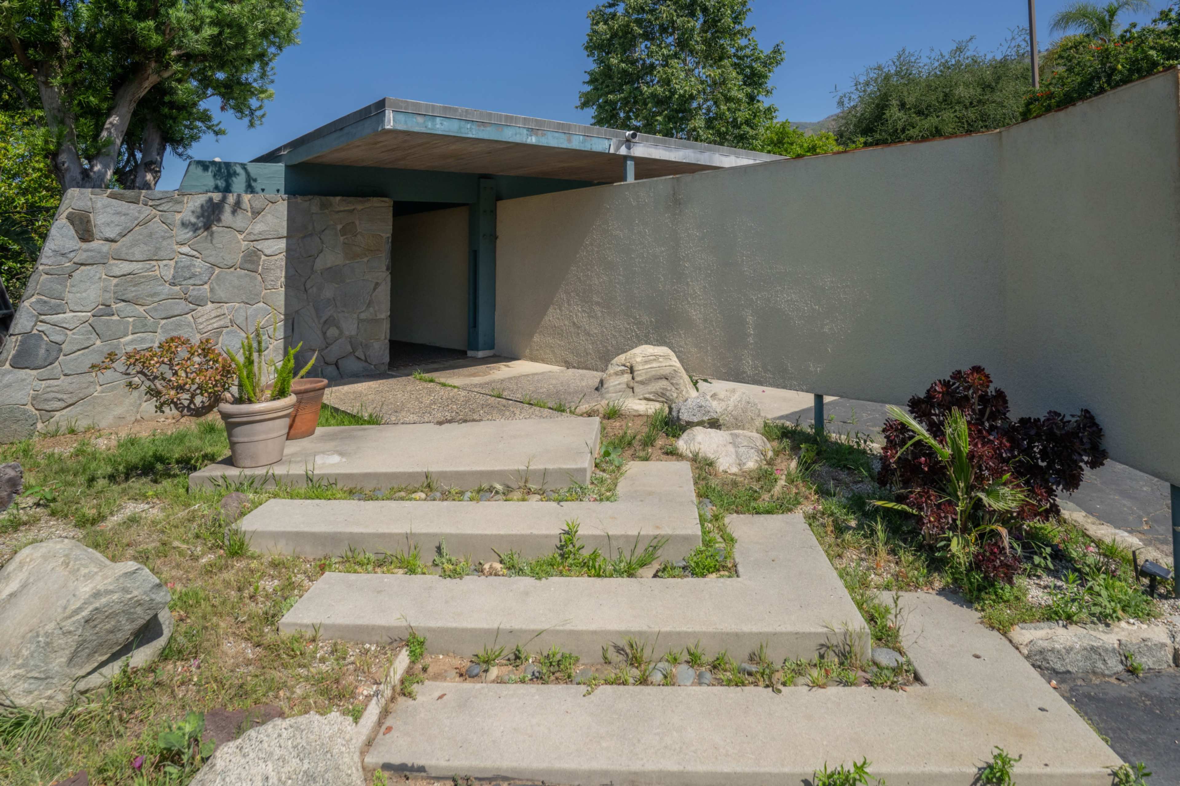 The image shows a modern entrance pathway leading to a building, flanked by rocks and potted plants.