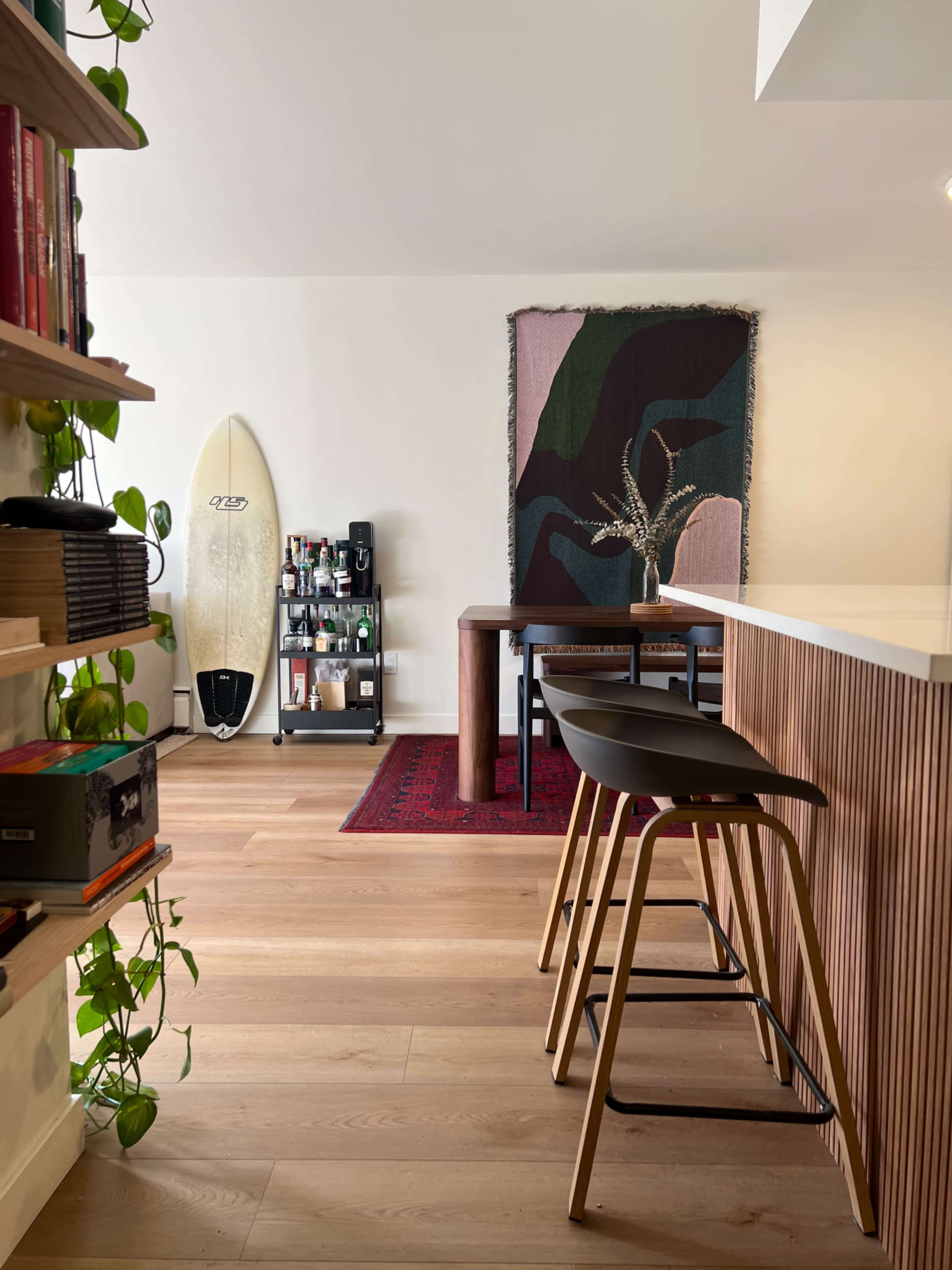 A modern living space featuring a surfboard against a wall, a wooden table with chairs, and a decorative rug.
