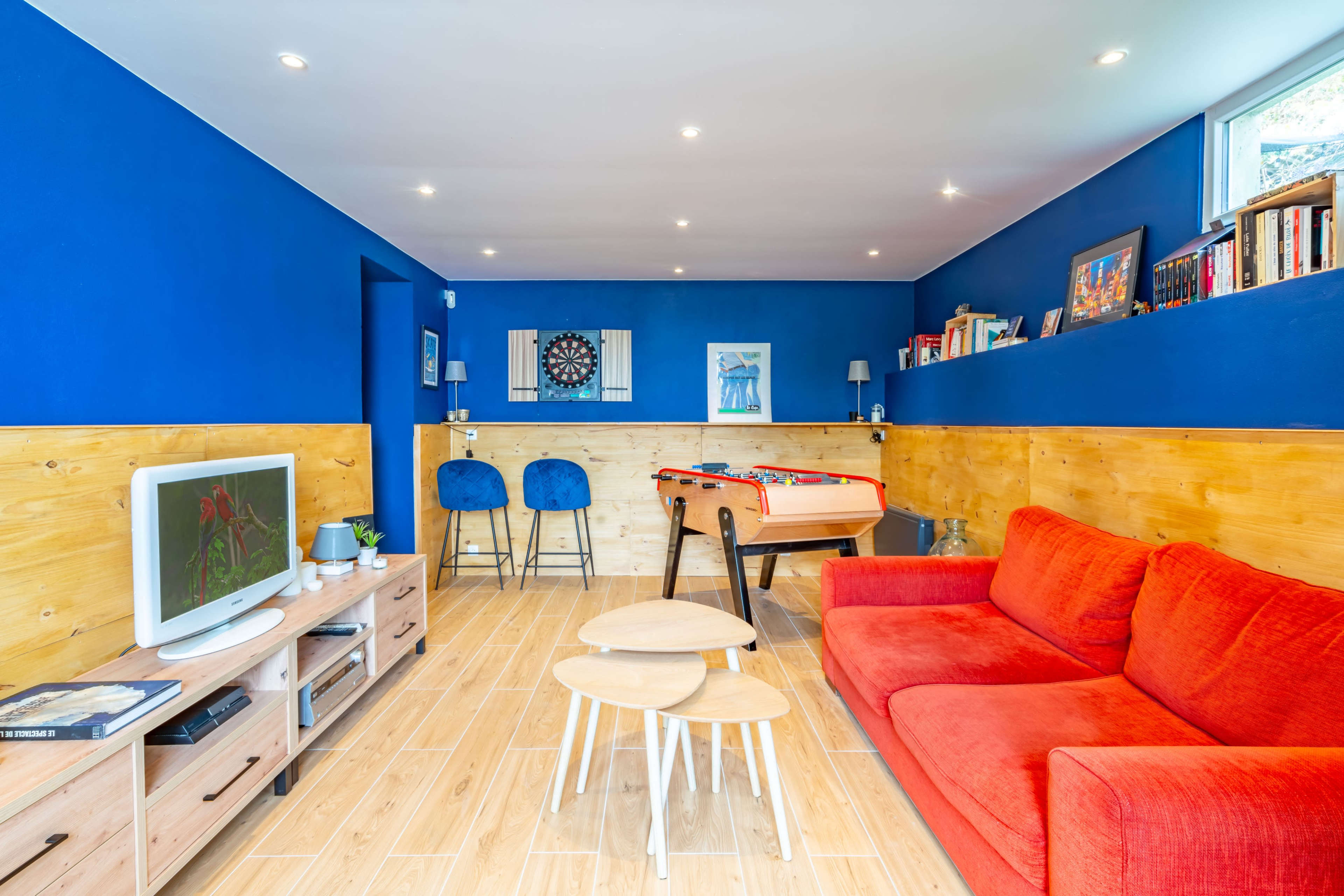 A modern room features a red sofa, a television, a foosball table, and blue bar stools against a blue wall with wooden paneling.