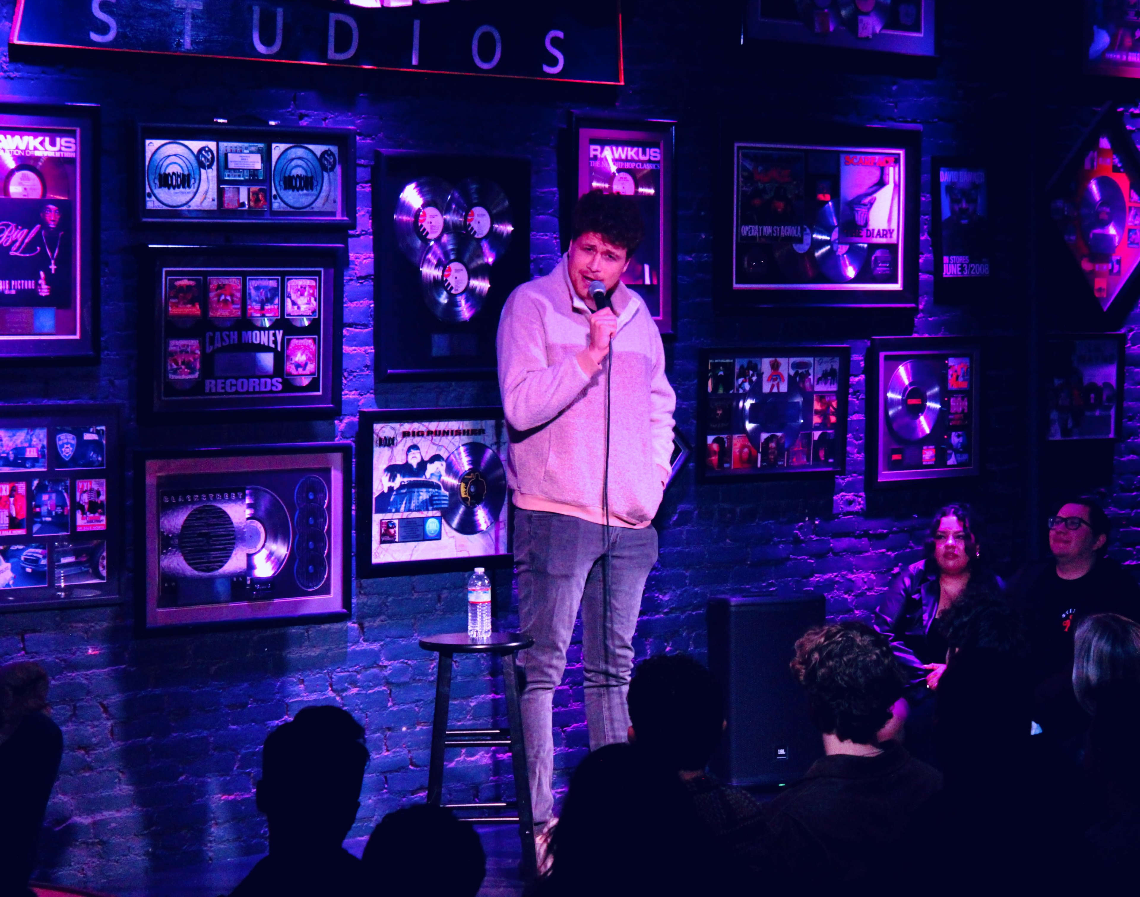 A comedian stands on stage with a microphone, performing in a venue adorned with framed music awards and records, while an audience watches.