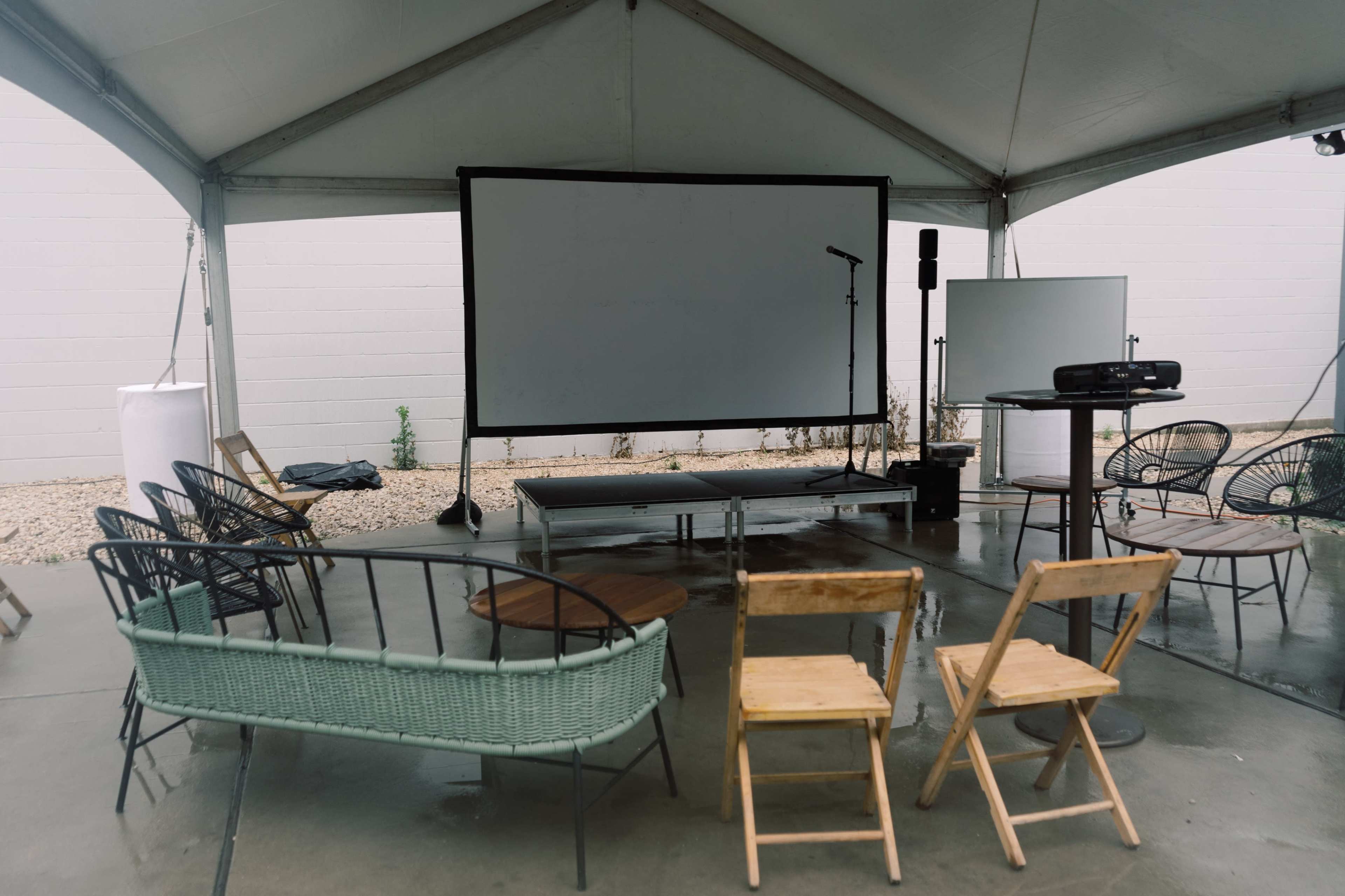 A tented area features a blank projection screen, a microphone on a stand, a projector, and various seating arrangements including chairs and a couch.