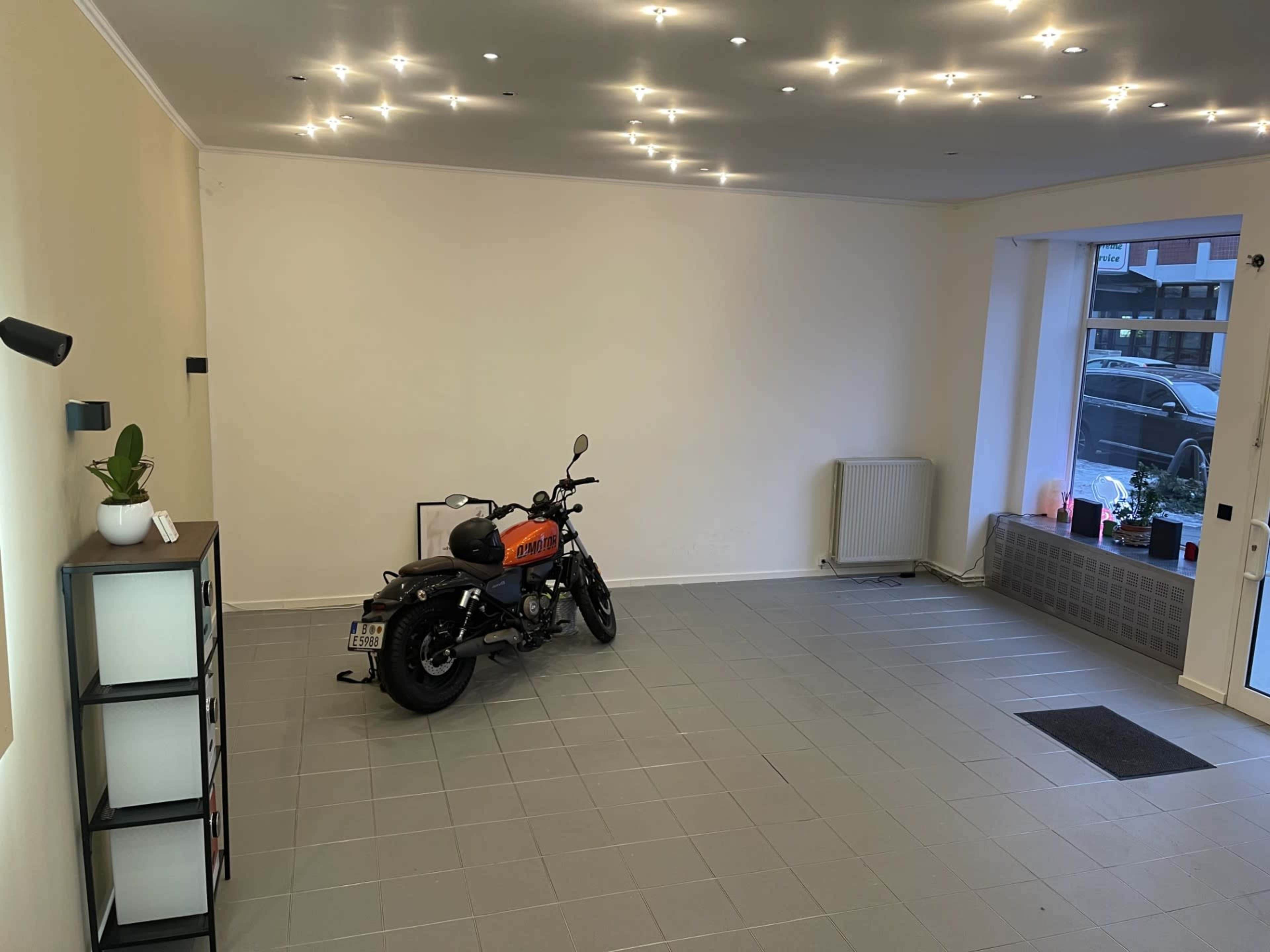 The image shows a spacious, well-lit room with a motorcycle positioned on the left side and a window on the right.