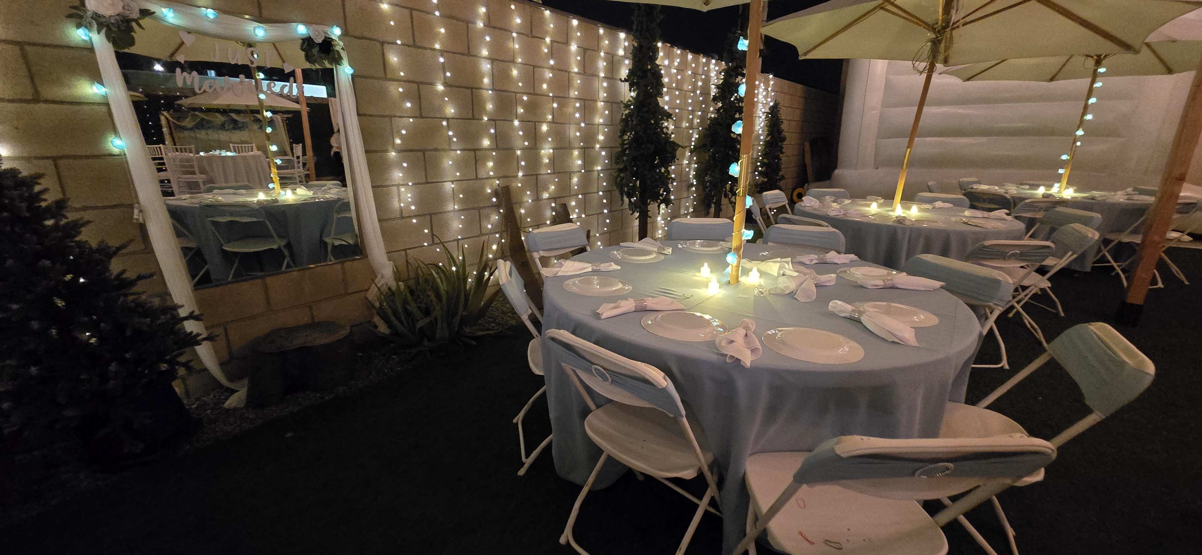 A dining area is set up outdoors with round tables covered in light blue tablecloths and illuminated by soft lighting from string lights.