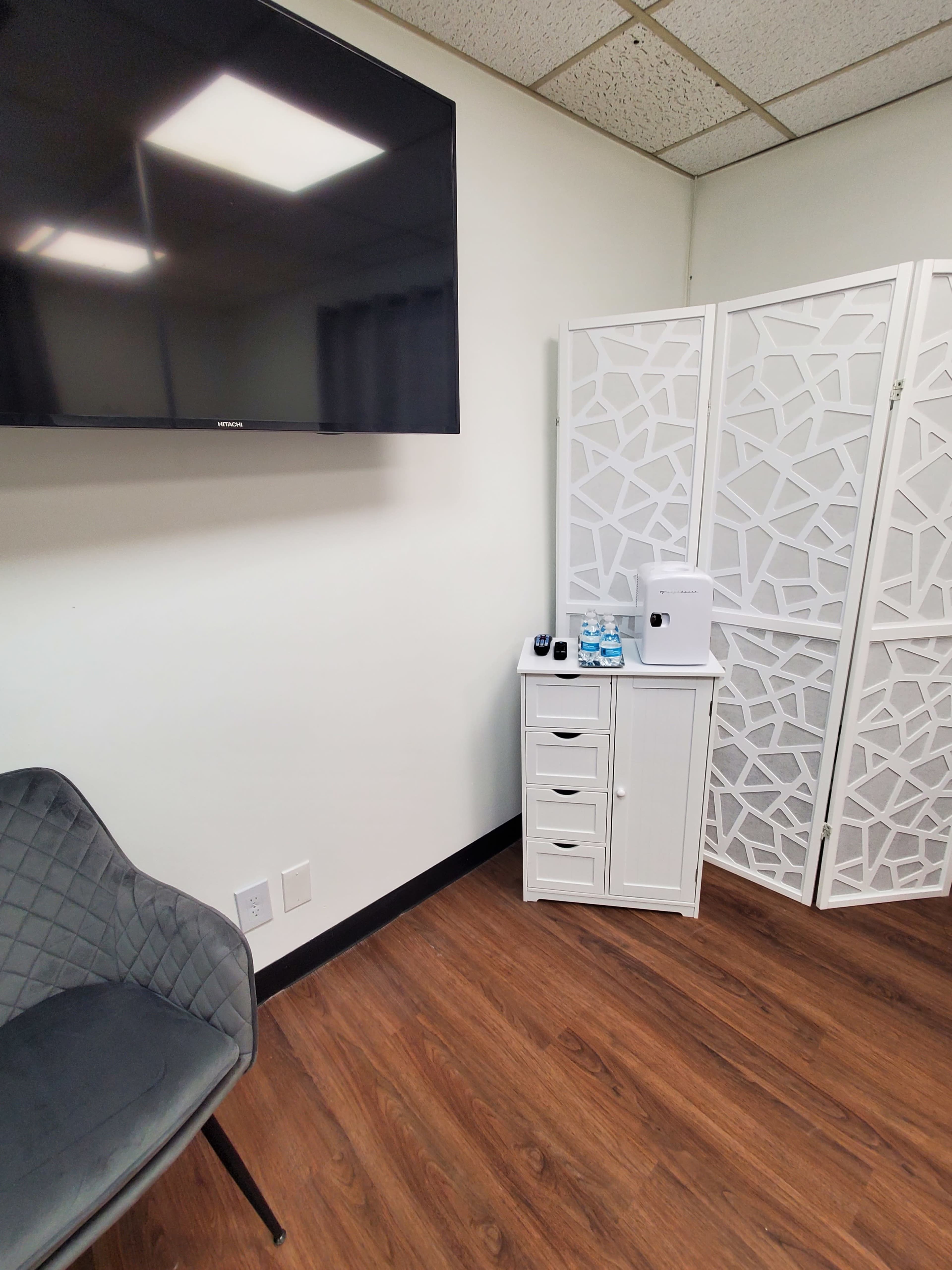 The image shows a small room with a TV mounted on the wall, a decorative partition, a white storage cabinet, and a gray chair on a wooden floor.
