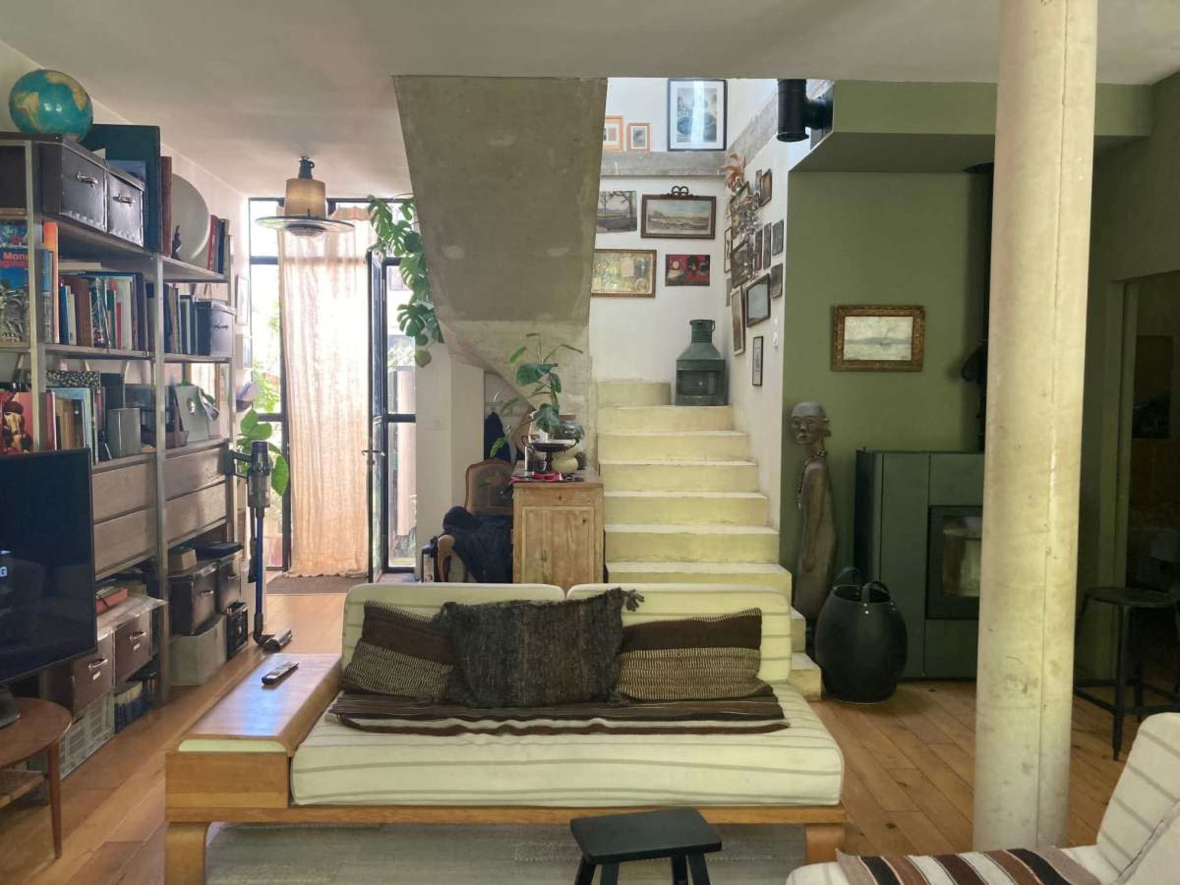 The image shows a modern living room with a staircase leading to an upper level, featuring a cozy sofa, bookshelves filled with books, and decorative items on the walls.