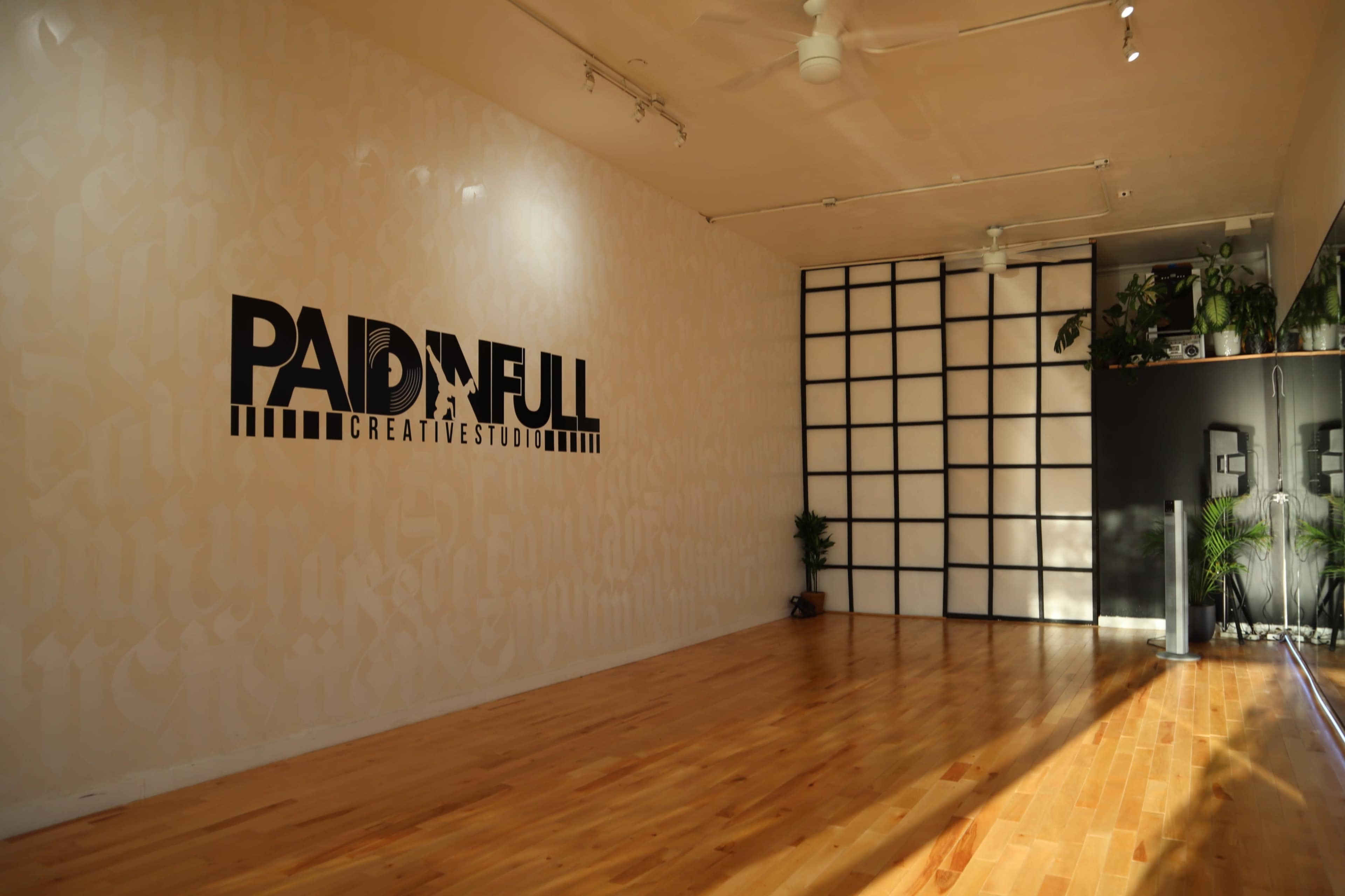 The image shows a well-lit studio space with a wooden floor, featuring a wall displaying the logo "PAID IN FULL CREATIVE STUDIO" and a grid-patterned wall on the right.