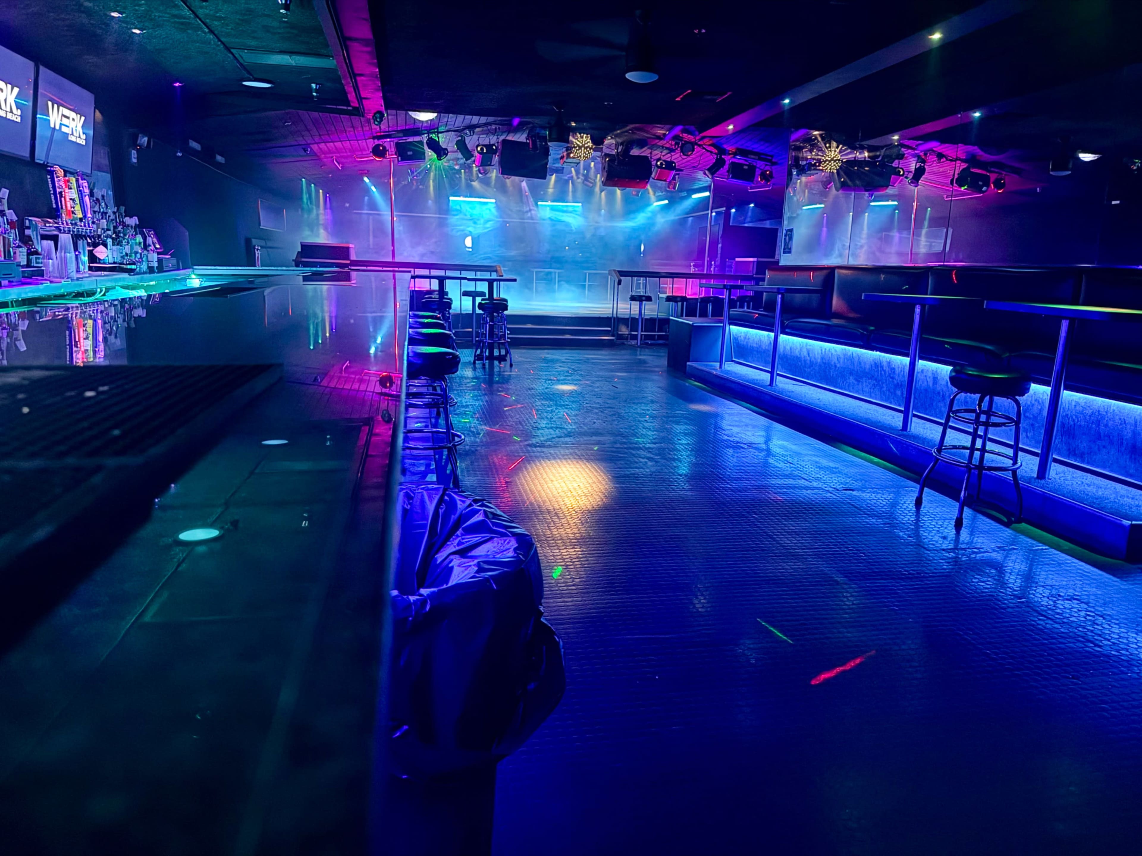 The image shows a dimly lit nightclub interior featuring a bar, scattered stools, and colorful ambient lighting effects on the dance floor.