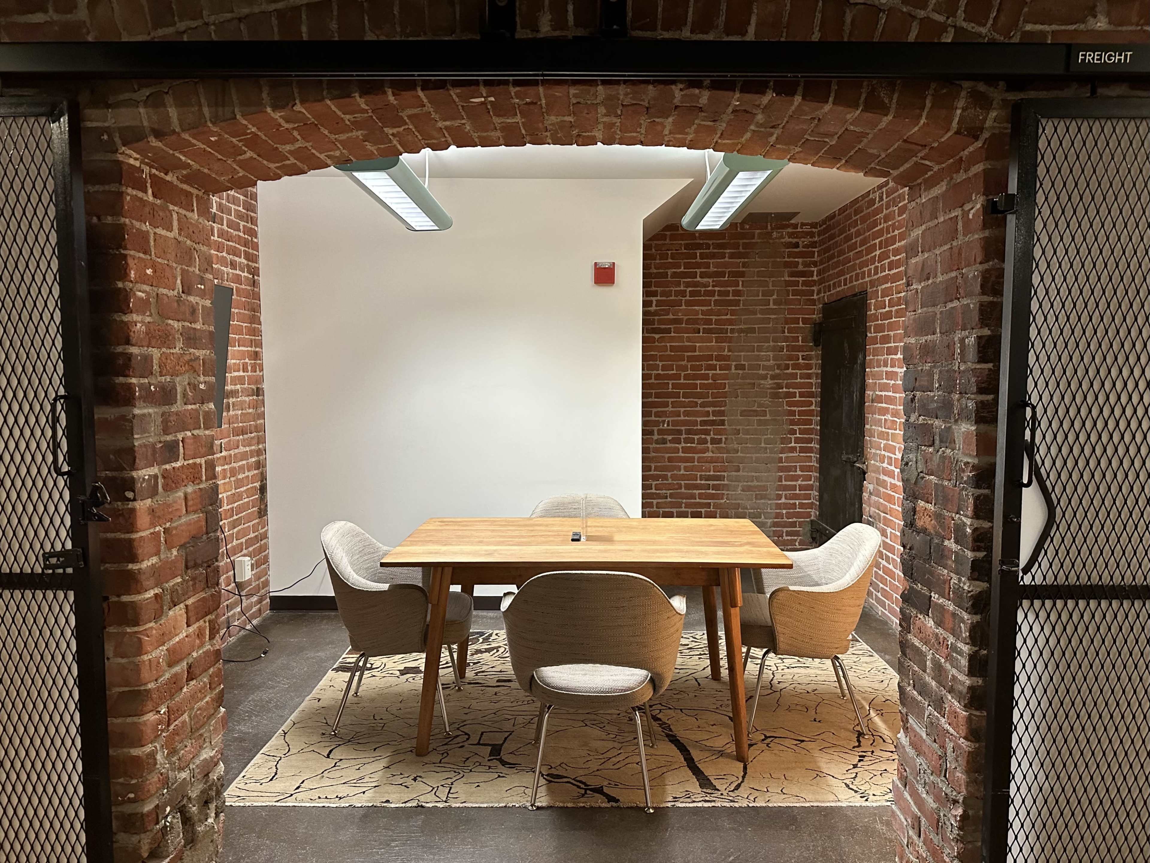 A brick-walled meeting room features a wooden table surrounded by four upholstered chairs on a patterned rug.